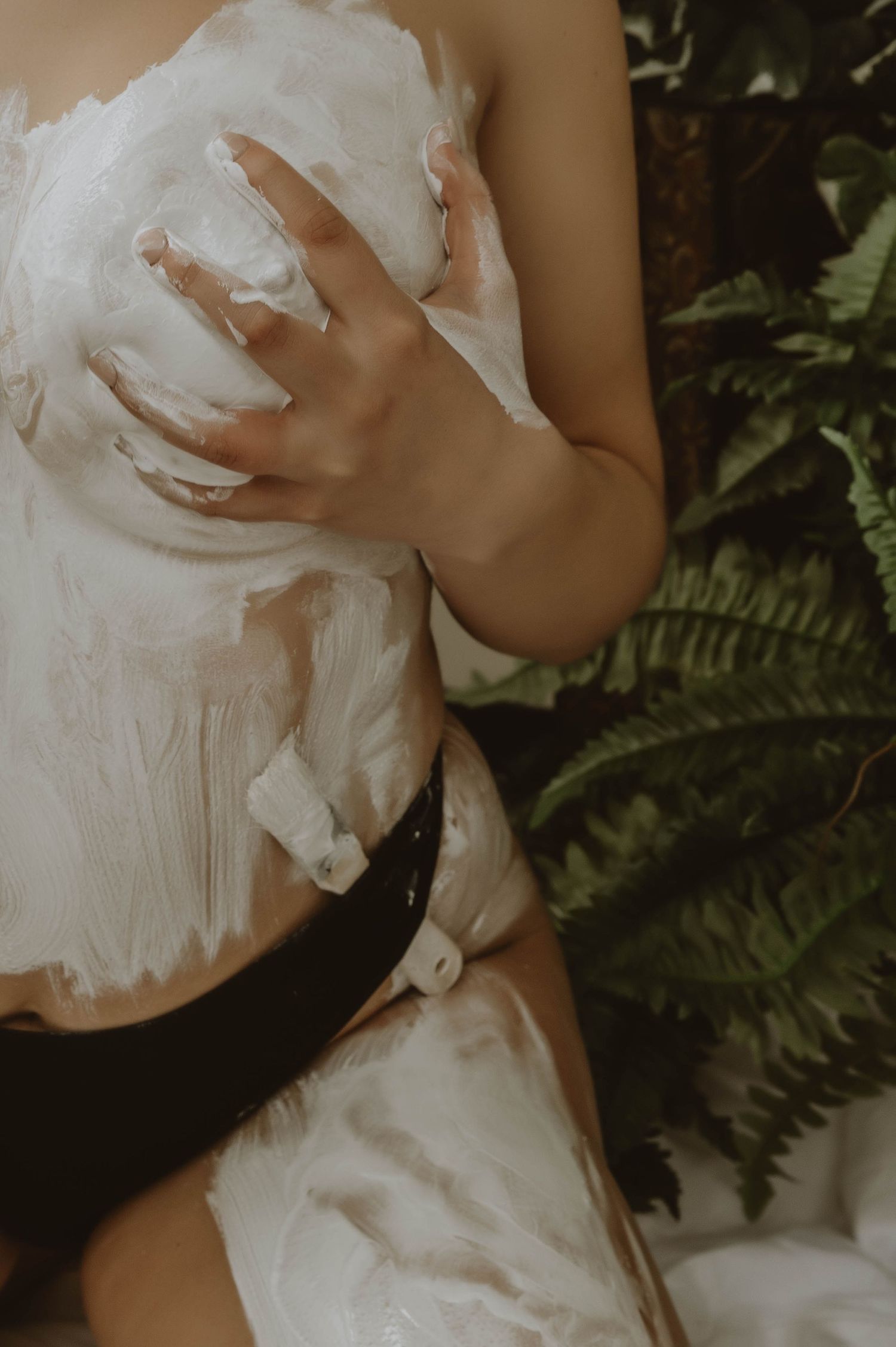 Cropped torso of a person covered in white cream or lotion, wearing black bottoms, posed beside green fern leaves in soft natural light.