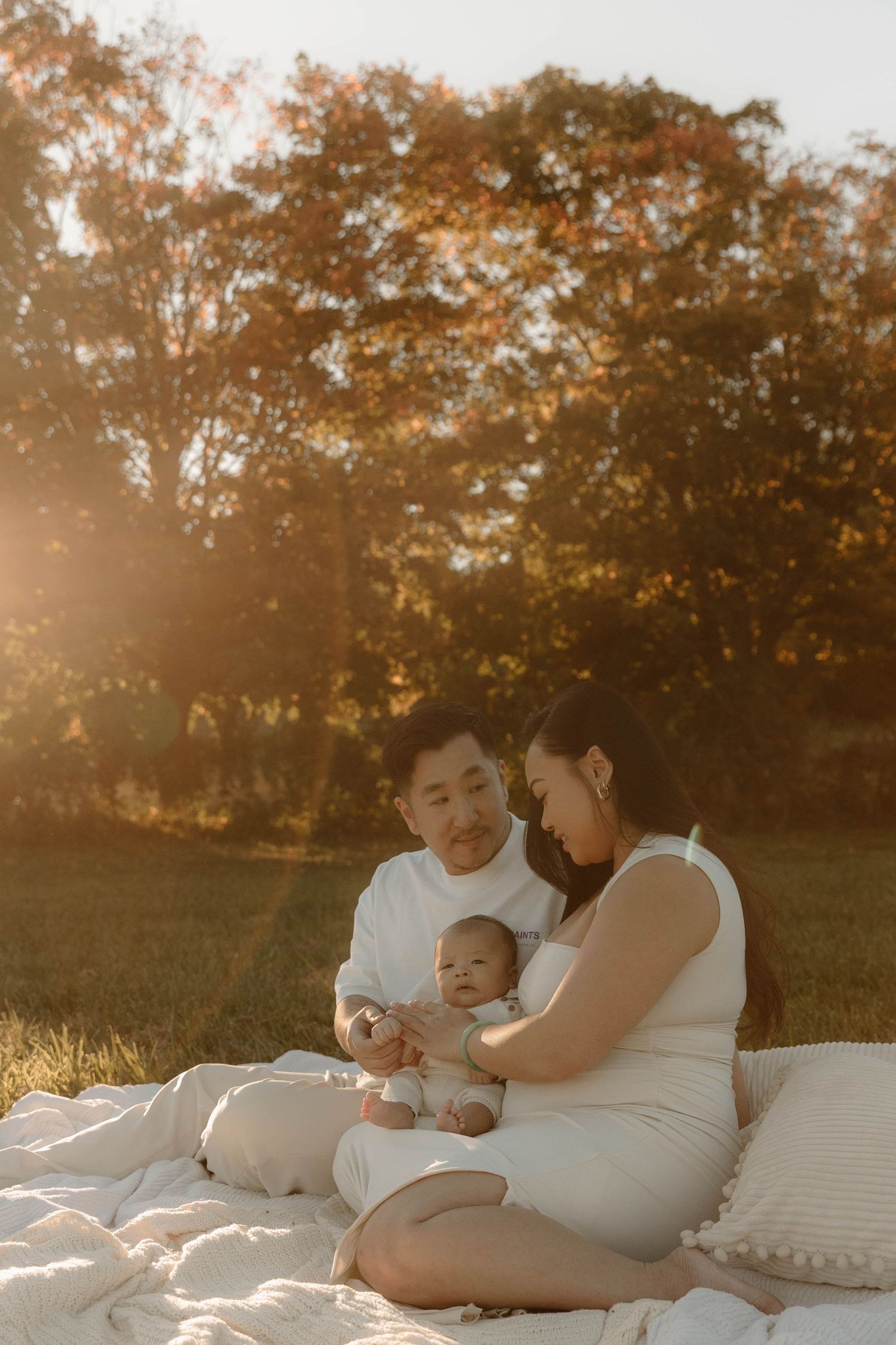 A family sits on a white blanket in a sunlit autumn park, parents lovingly gazing at their baby.