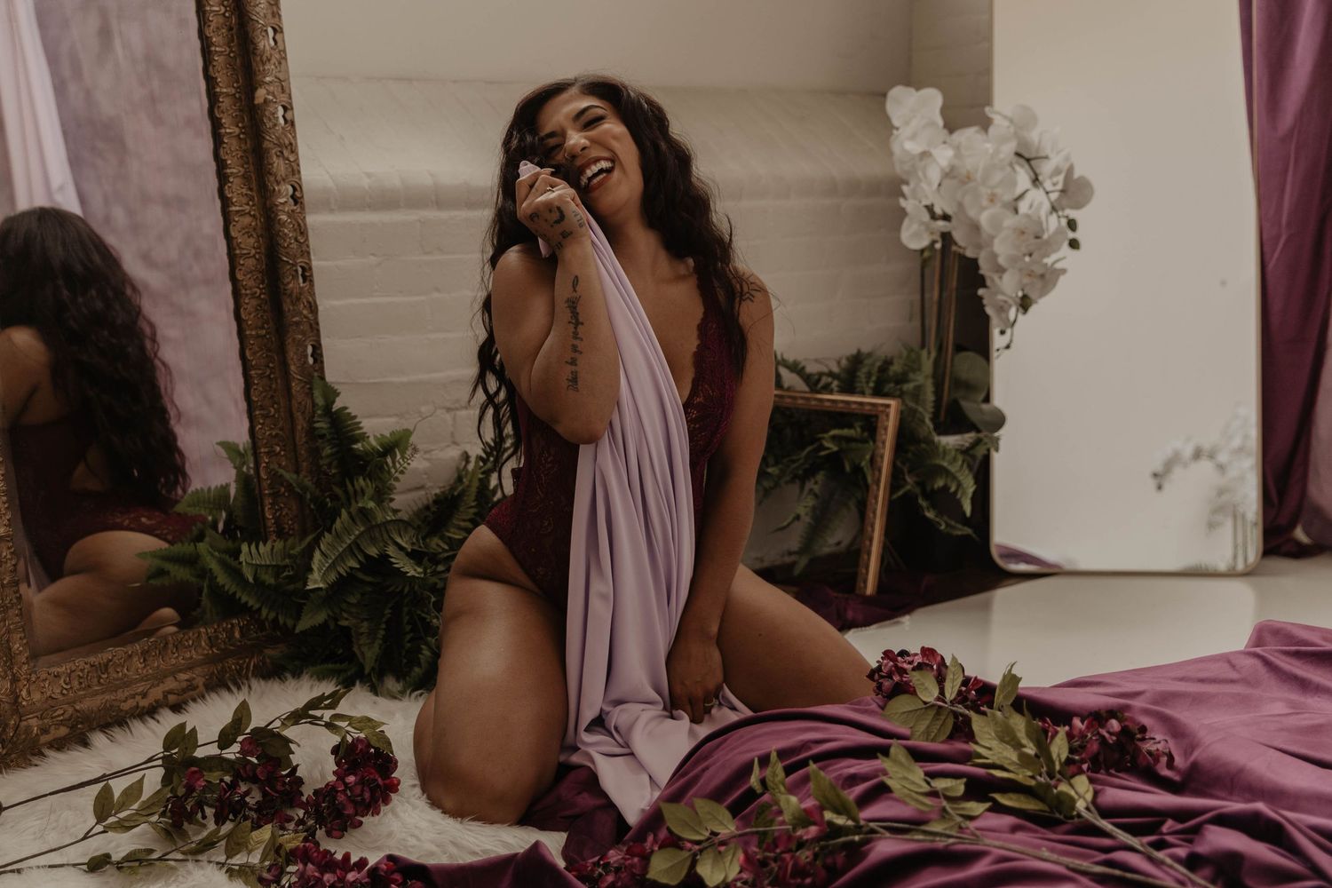 Smiling woman with long dark hair sits on the floor in a burgundy bodysuit, wrapped in a mauve drape, surrounded by flowers and a mirror.