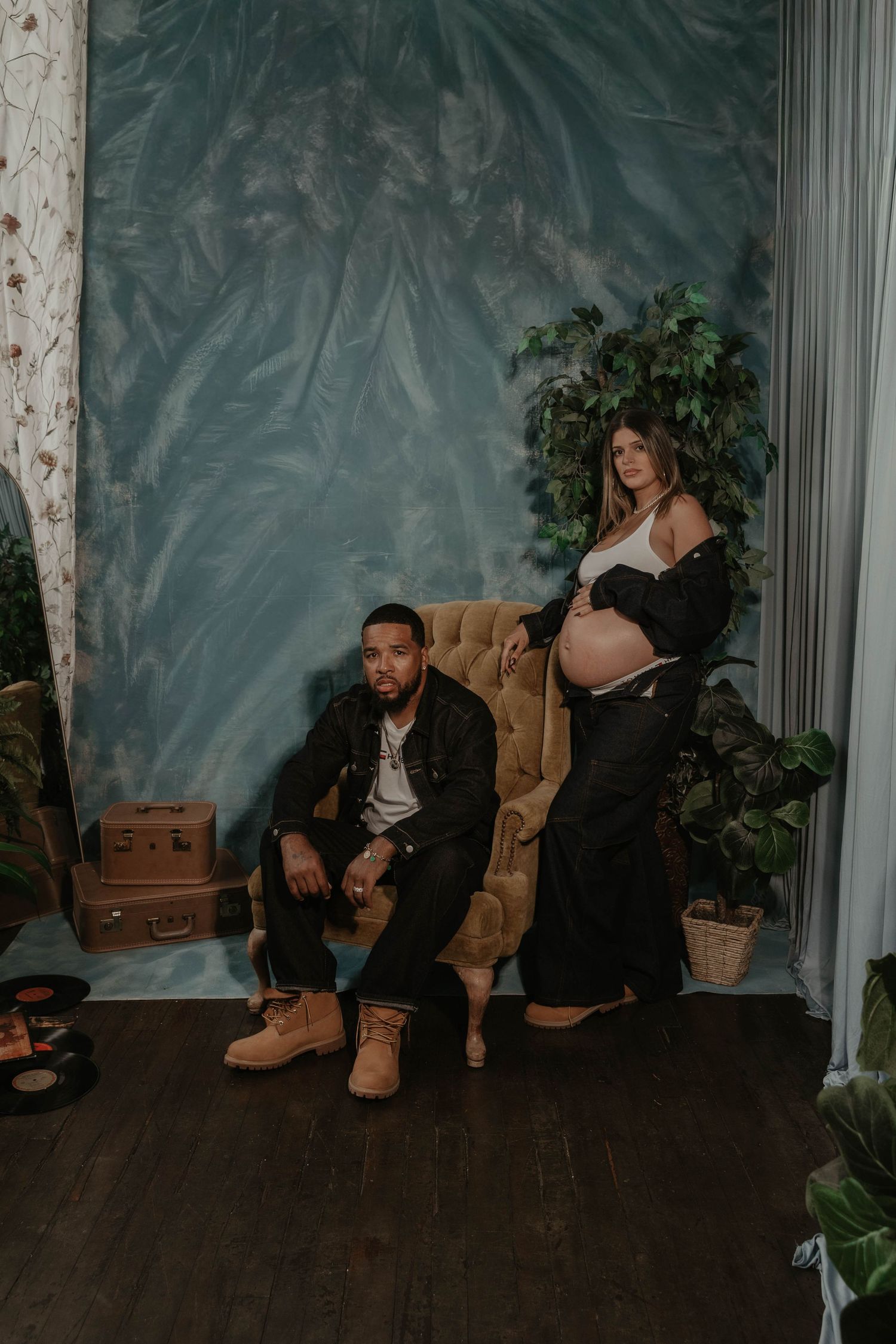 A couple poses for a maternity photo shoot, man seated in velvet chair, pregnant woman standing beside him in a studio.