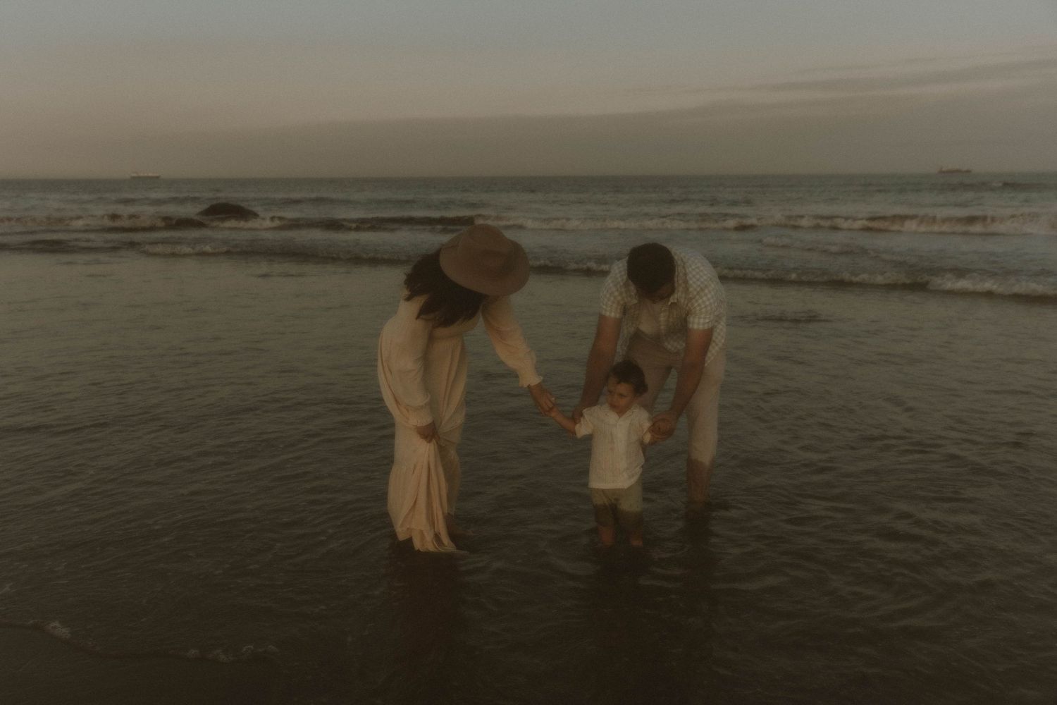 Family of three holding toddler hands at ocean shoreline during golden hour sunset.