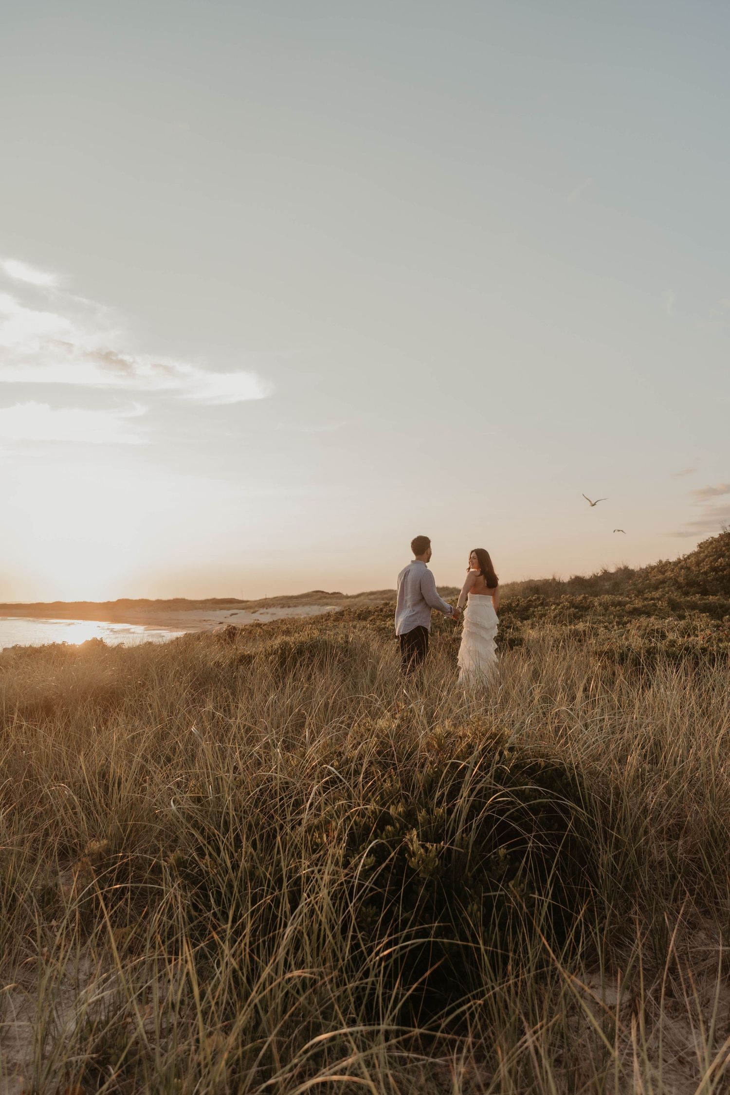 Couple holding hands in golden sunset light among coastal dunes, woman in white dress, romantic beach engagement photo.
