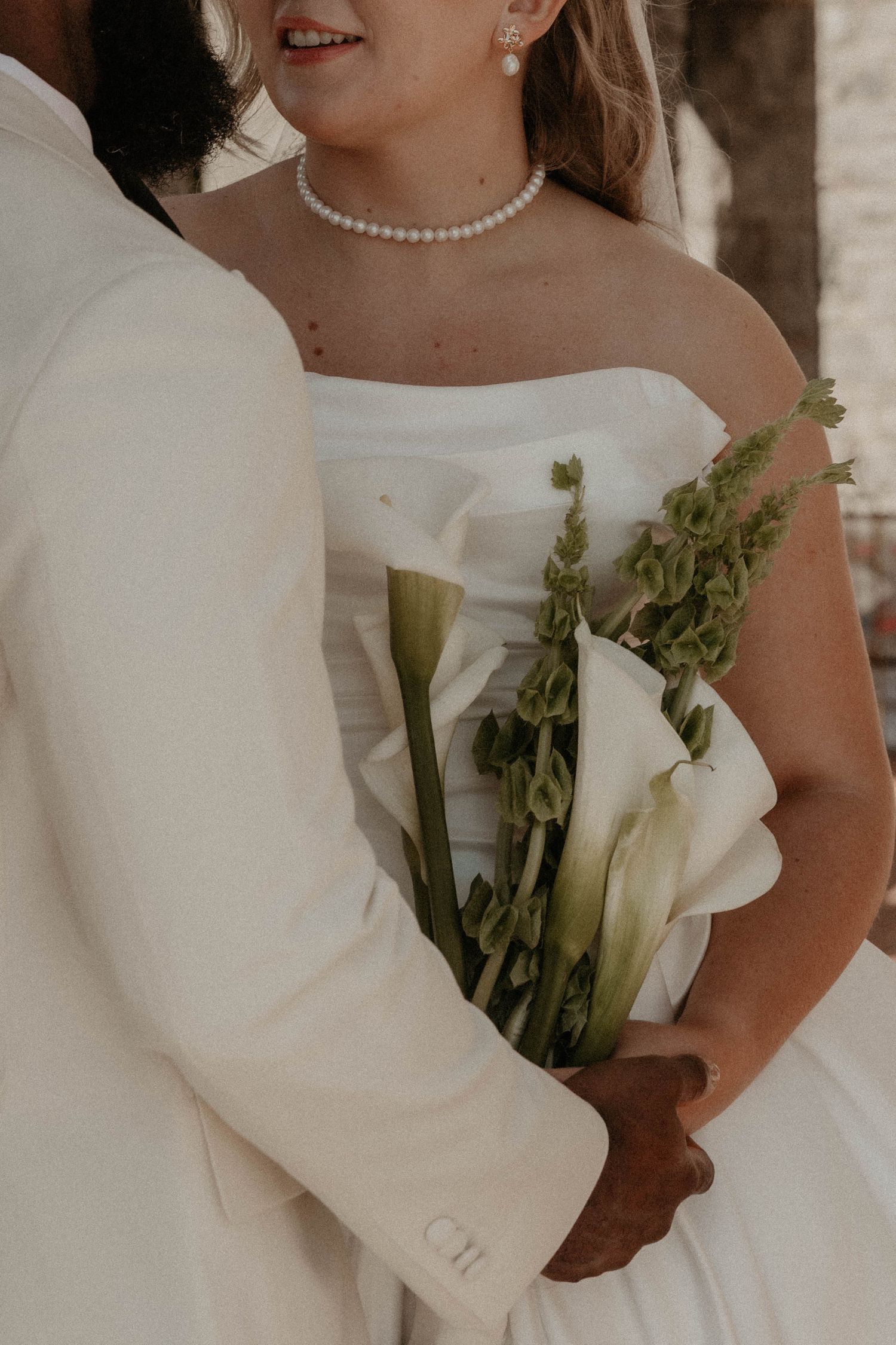 Bride in strapless white gown holding calla lily bouquet with groom in cream suit at elegant wedding ceremony.