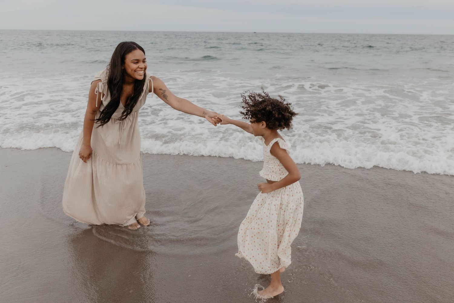 A smiling woman in a flowing beige dress holds hands with a young girl in a white dress as they play in the ocean waves on the beach.