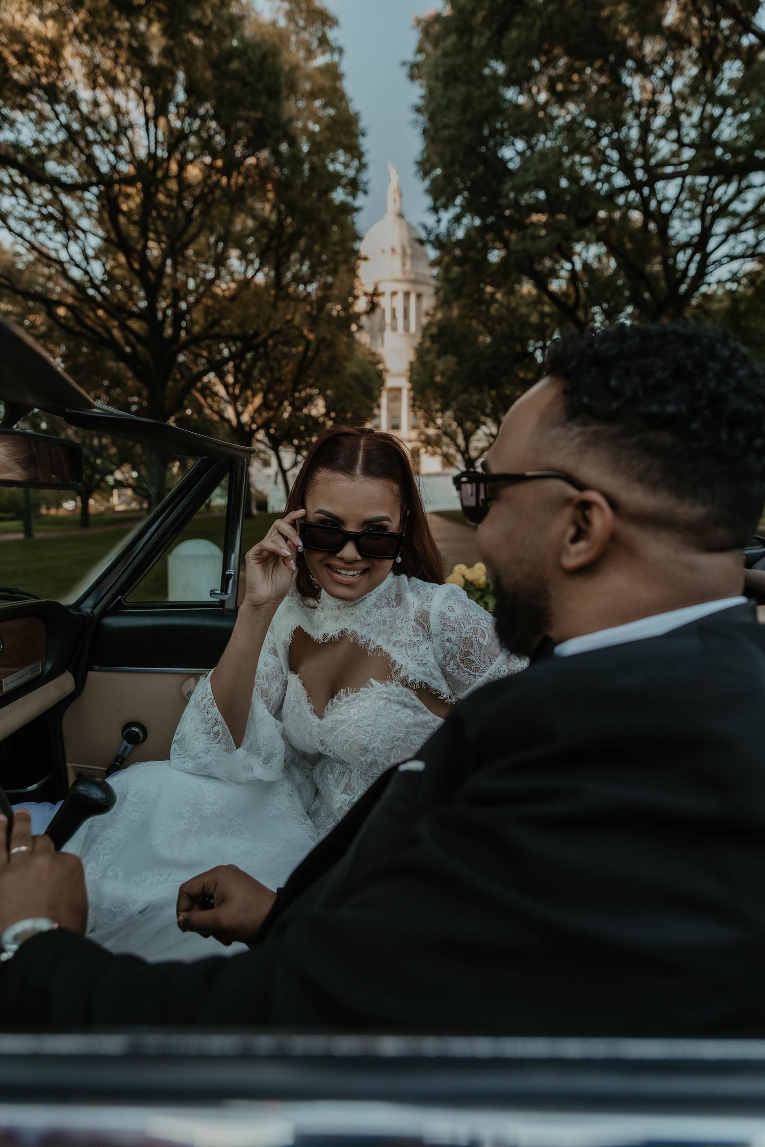 Smiling bride in a lace wedding gown lowers her sunglasses while seated in a vintage convertible beside her groom near a domed capitol.