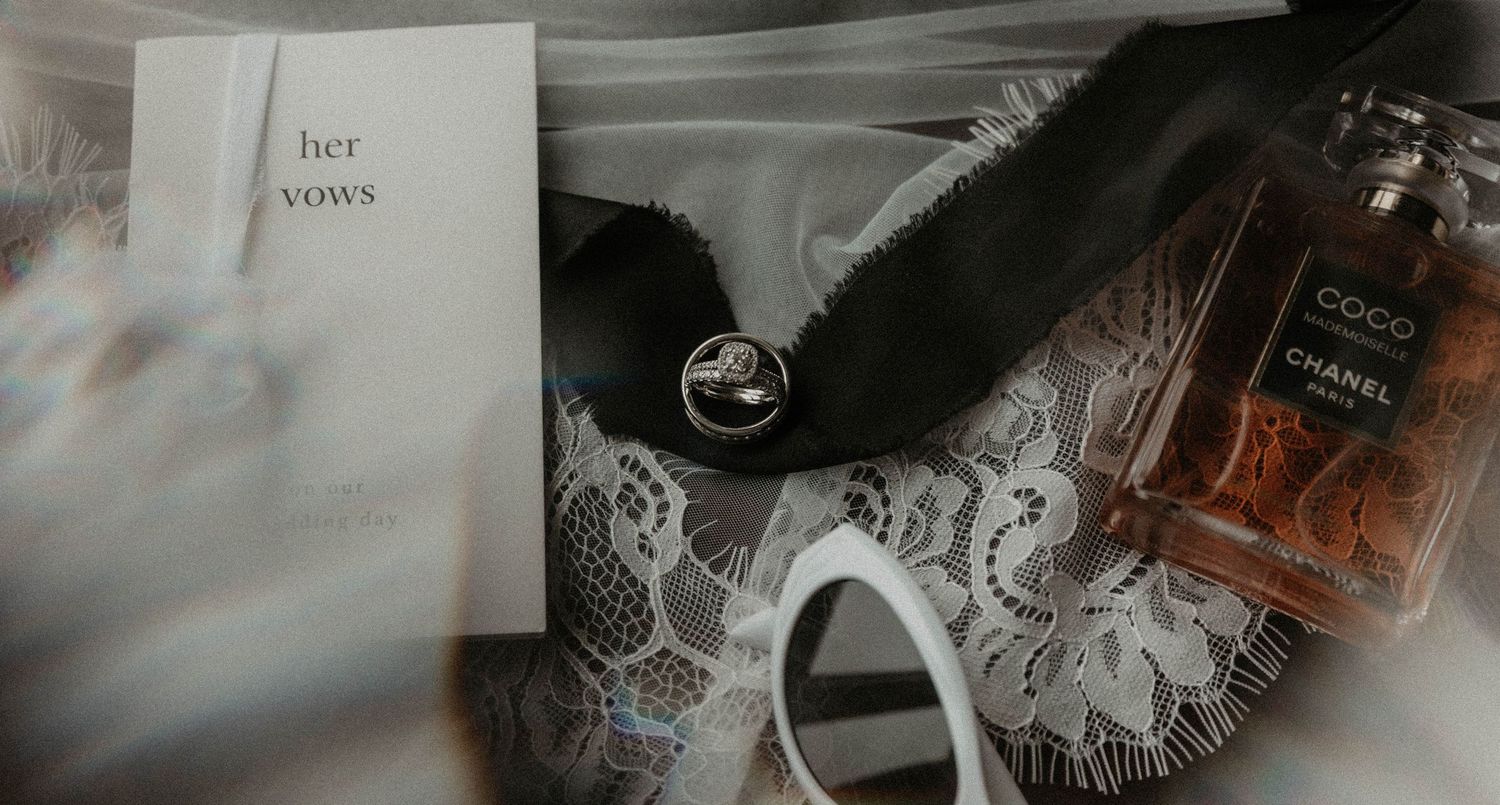 Wedding flat lay with vows card, rings, black feather mask, lace, and Chanel Coco perfume bottle.