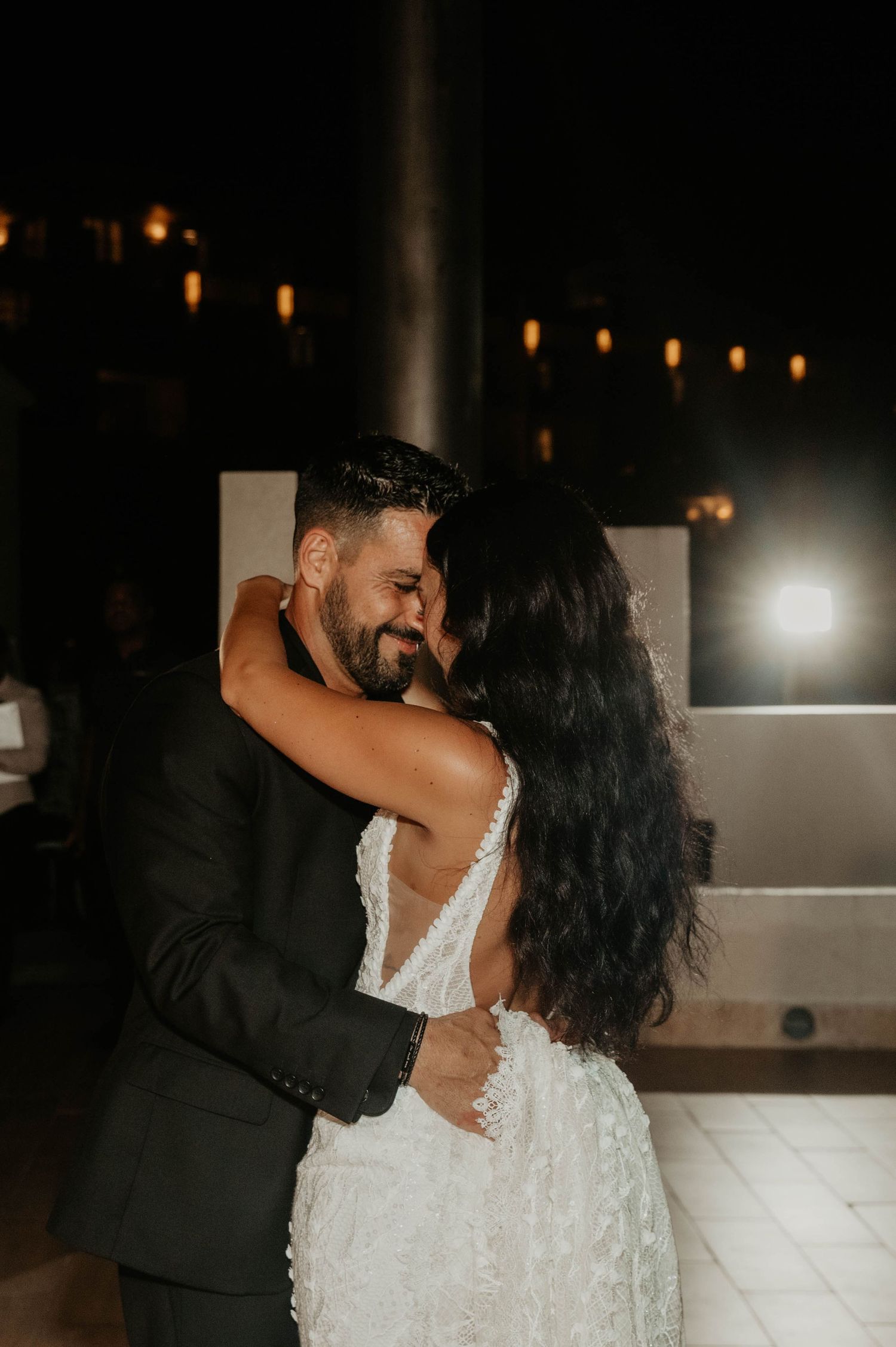 Bride and groom share an intimate first dance embrace at night, glowing string lights in the background.