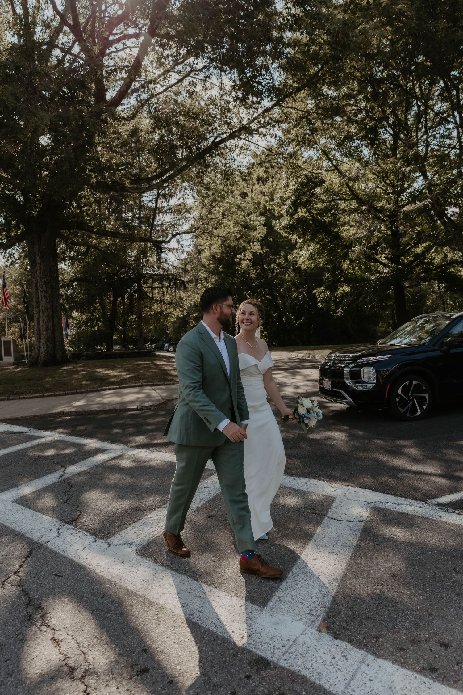 Bride and groom crossing a street hand in hand, bride in white gown, groom in sage suit, surrounded by lush trees.