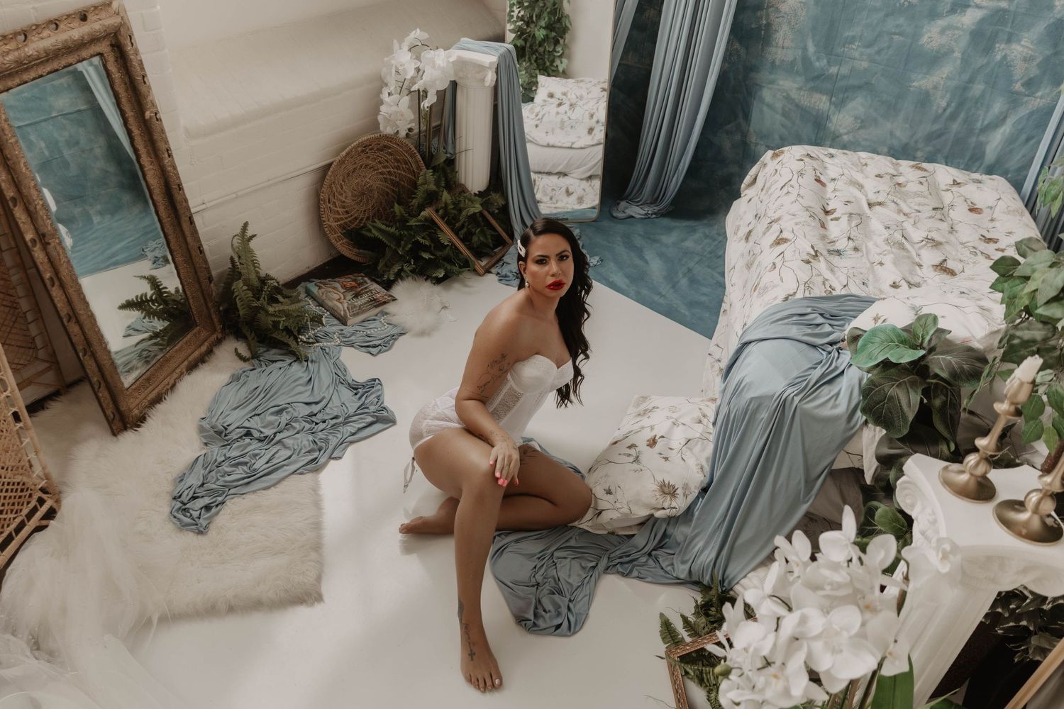 Woman in white lingerie poses on floor surrounded by lush greenery, floral decor, and soft blue drapery in elegant studio.