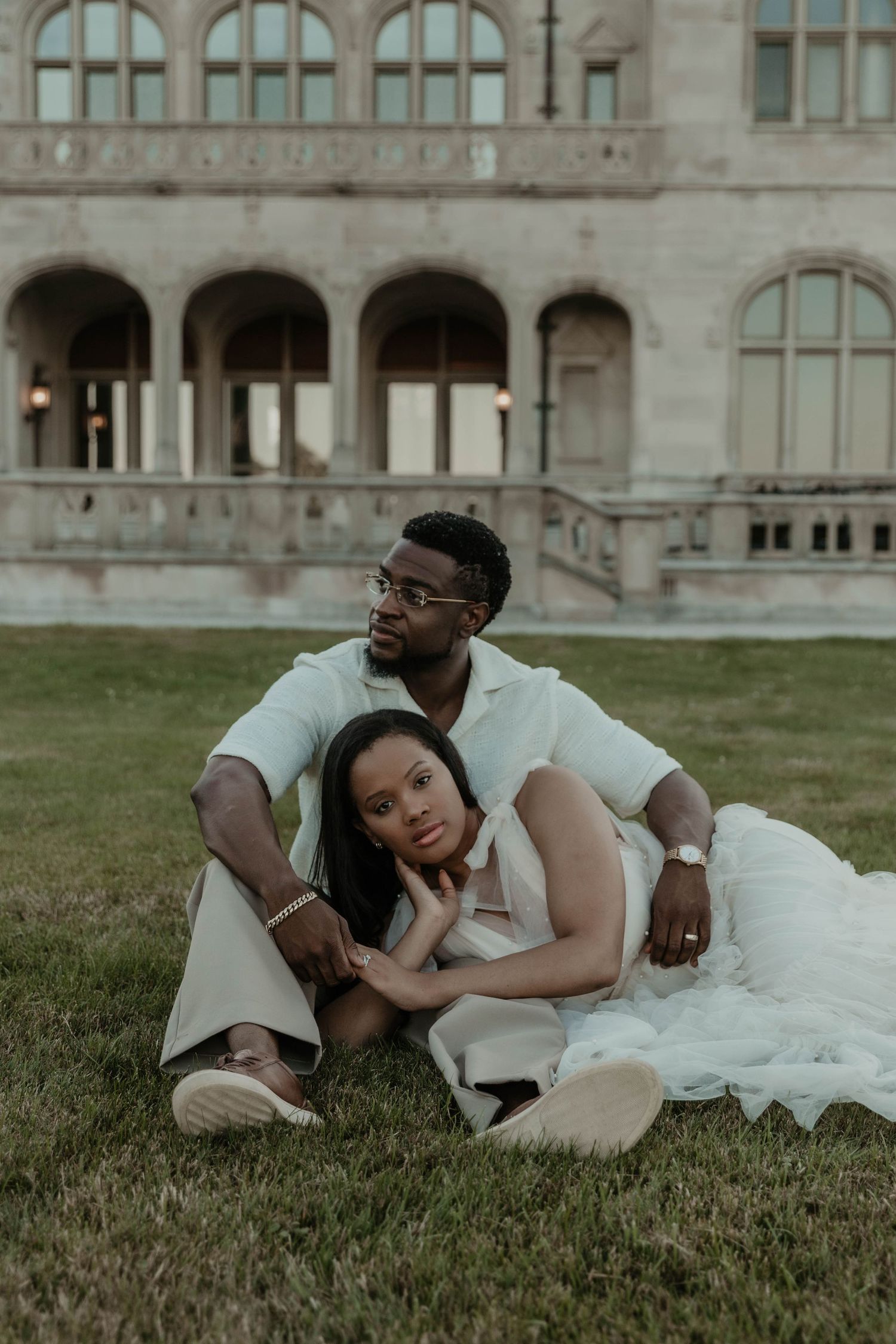 A couple in elegant white outfits sits on grass before a grand stone building, sharing a tender romantic moment.