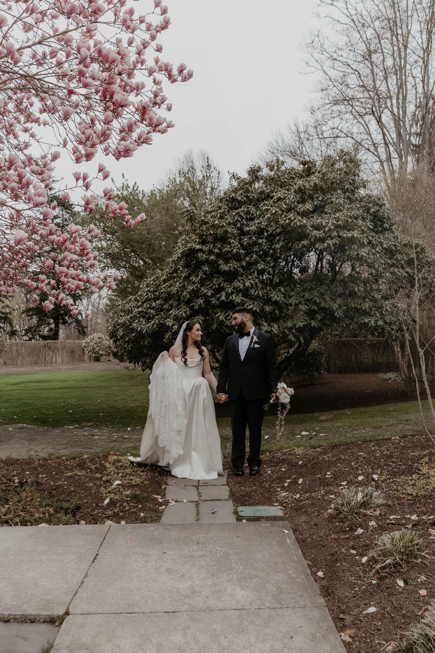 Bride and groom walk hand-in-hand on a garden path beside a blooming pink magnolia tree on their wedding day.