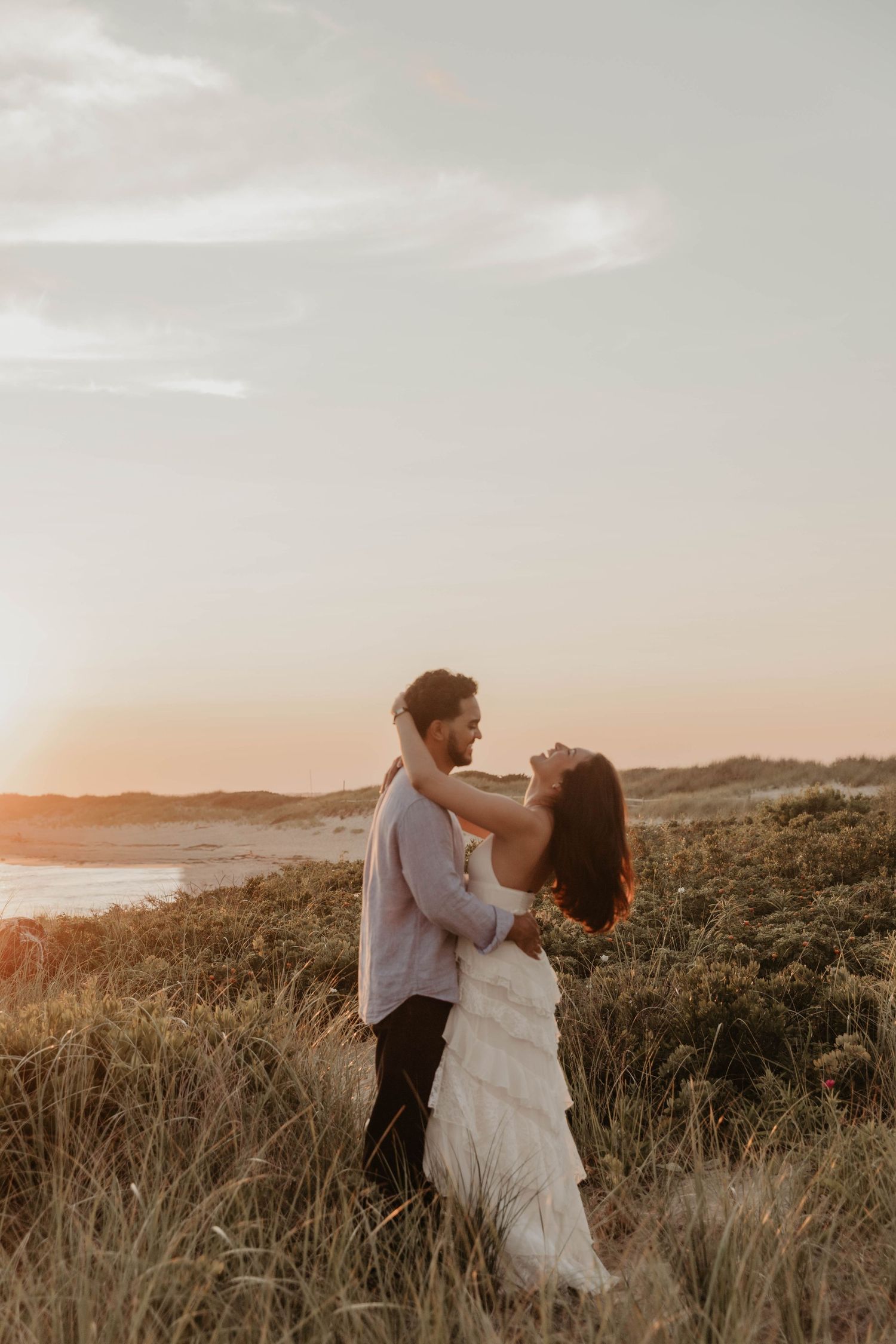 Couple embracing in golden sunset light among coastal grasses, bride in white tiered gown, romantic beach elopement photo.