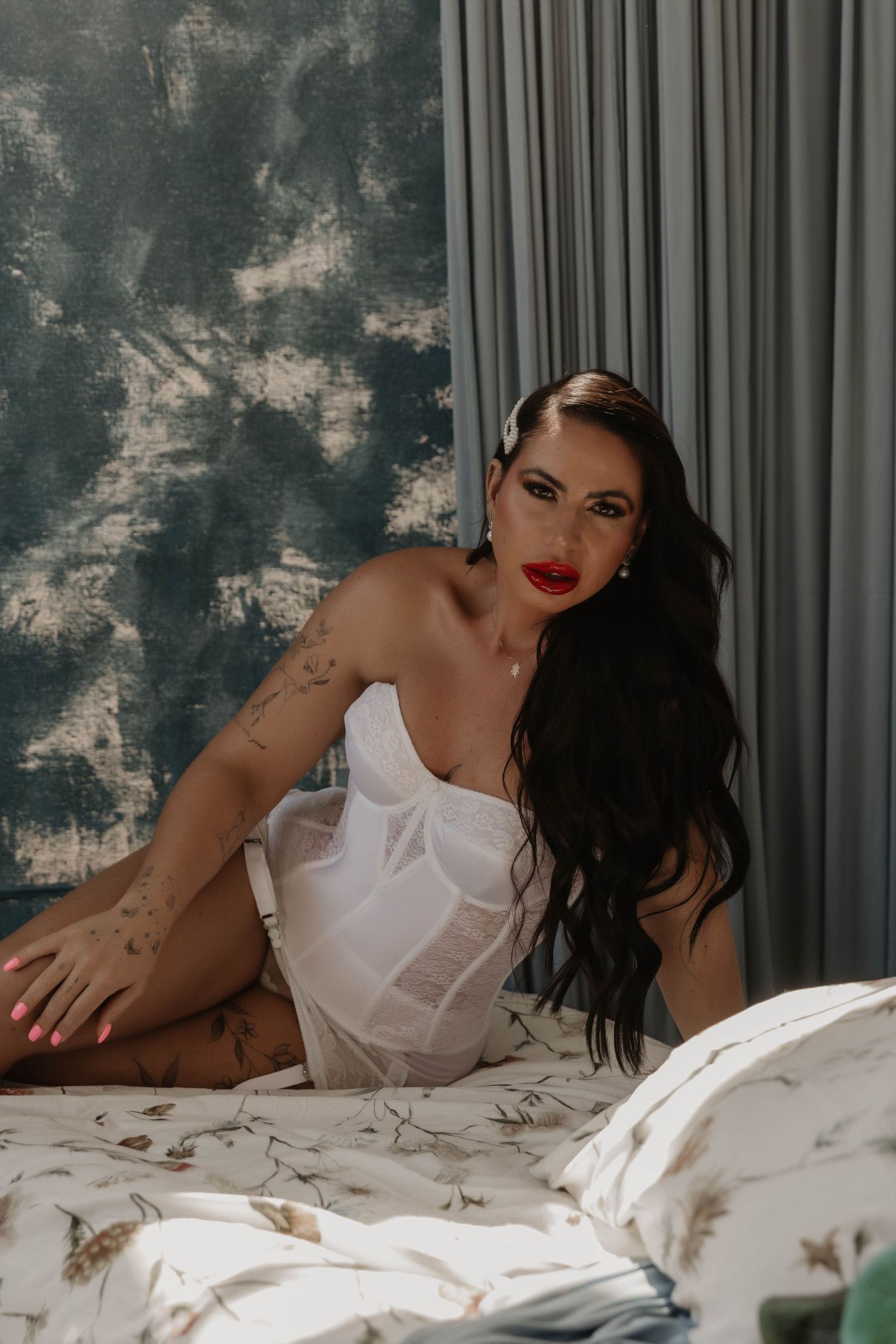 A woman with long dark hair and red lips poses in a white corset on a bed with floral bedding.