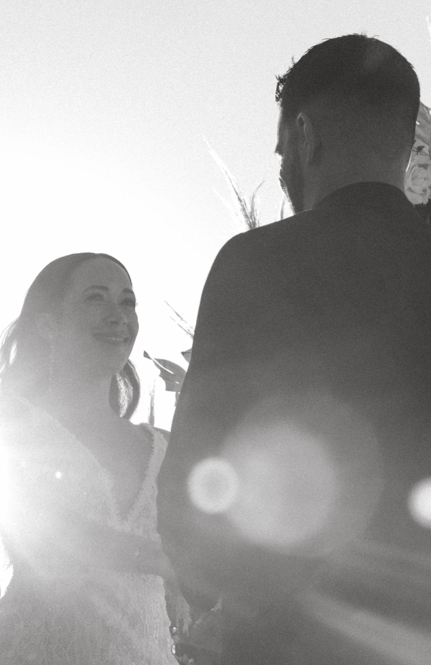 Black and white wedding ceremony photo with sun flare, bride smiling at groom outdoors.