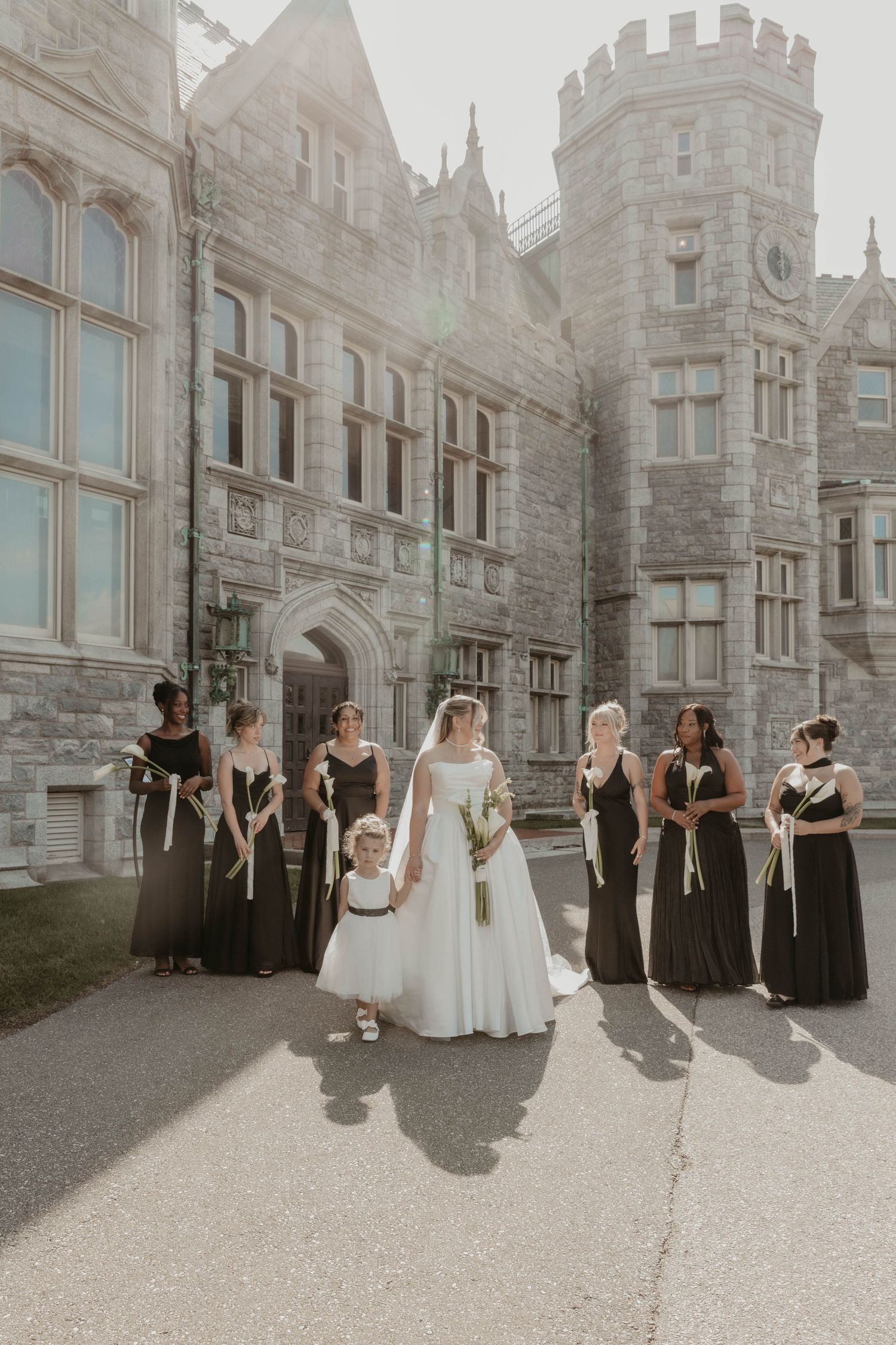 Bride in white gown with bridesmaids in black dresses posing outside a grand stone castle venue.