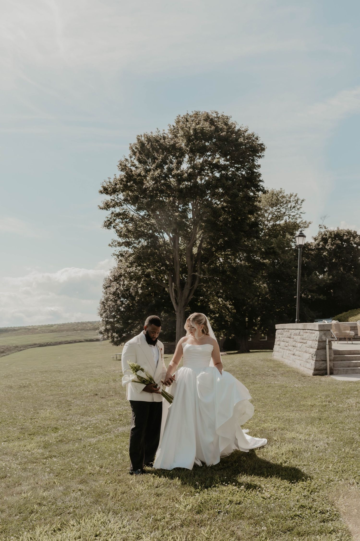 Bride and groom walking hand in hand on green lawn outdoors, bride in white ball gown, groom in white jacket, large tree behind.