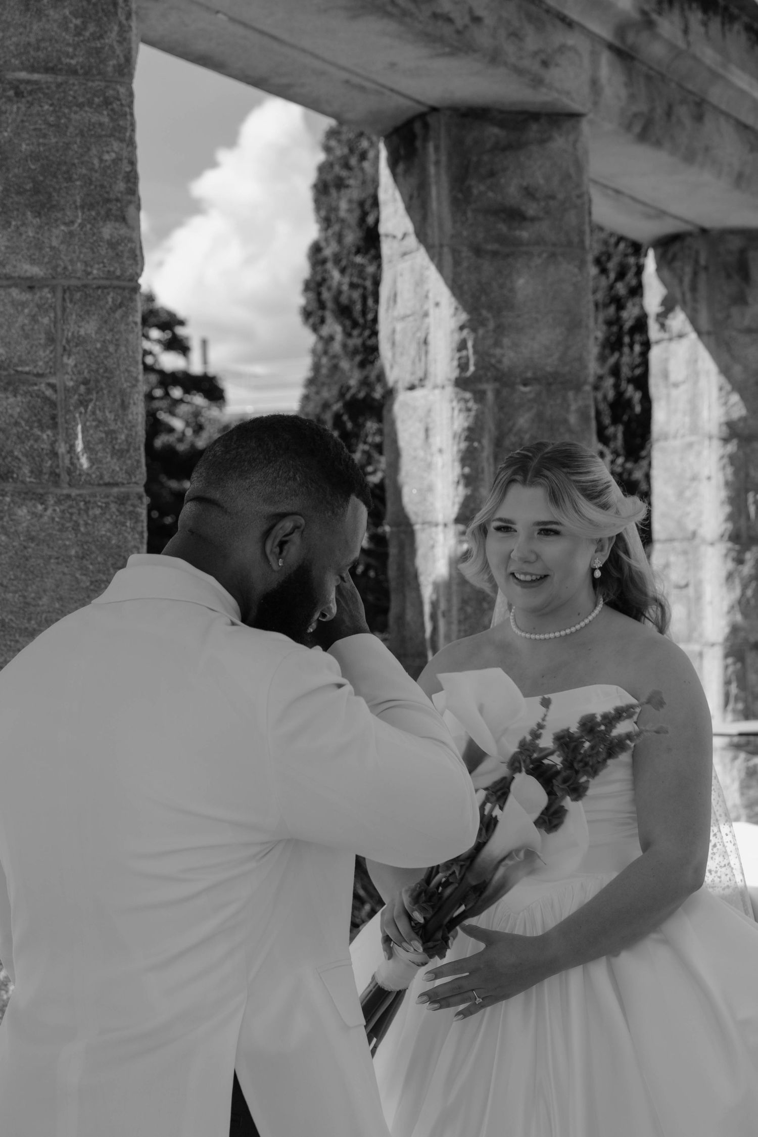 Black and white wedding photo of a smiling bride in white gown holding bouquet with groom in white suit.
