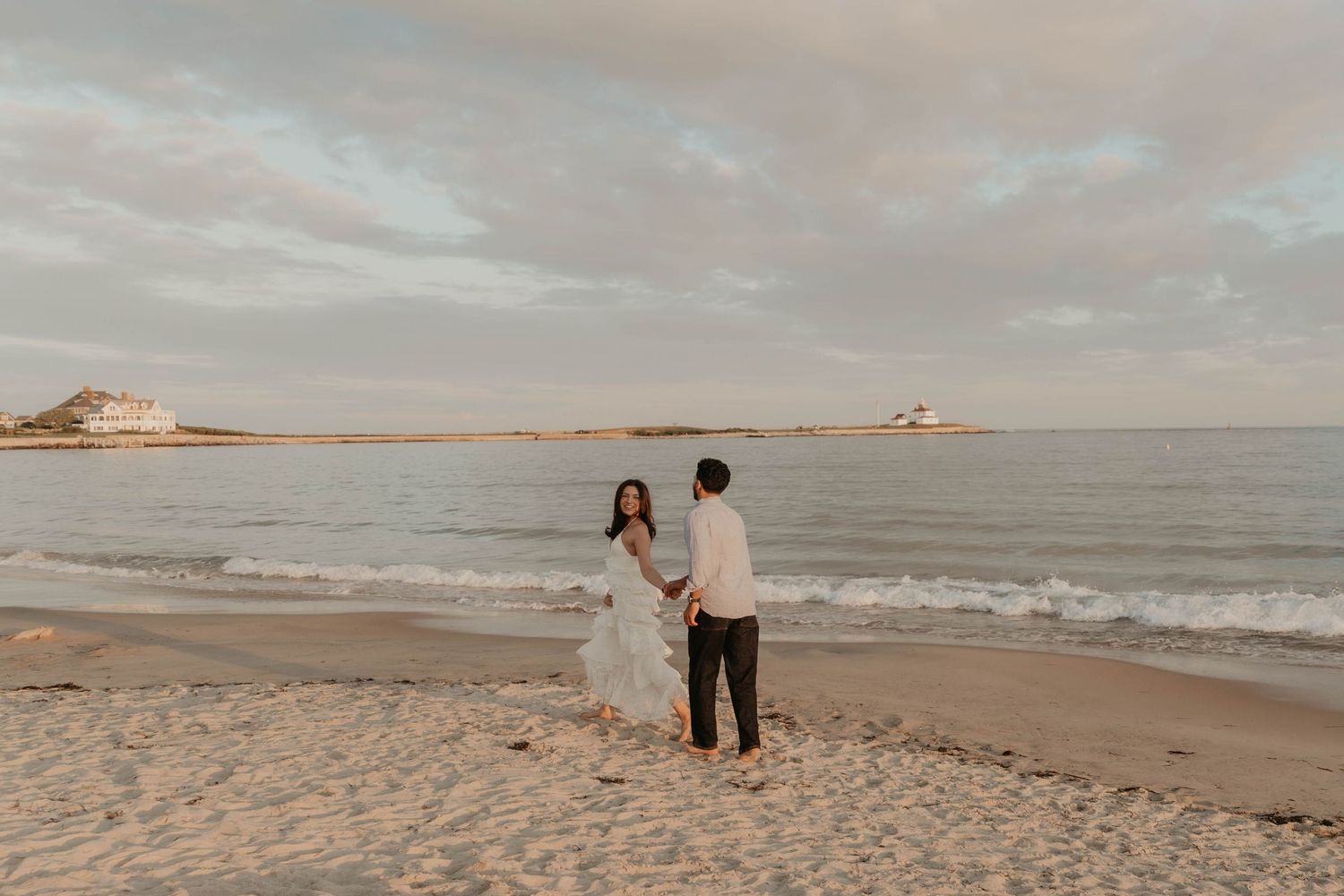 Couple holding hands on a sandy beach at sunset with calm ocean waves and a lighthouse in the background.
