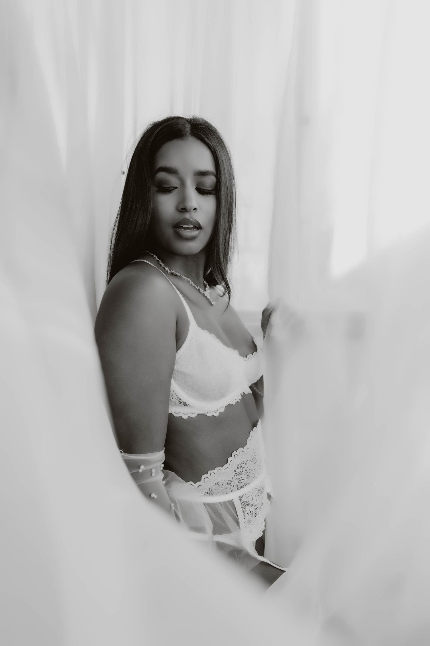 Black and white boudoir portrait of a woman in a lace bralette and sheer gloves, standing among flowing white curtains.