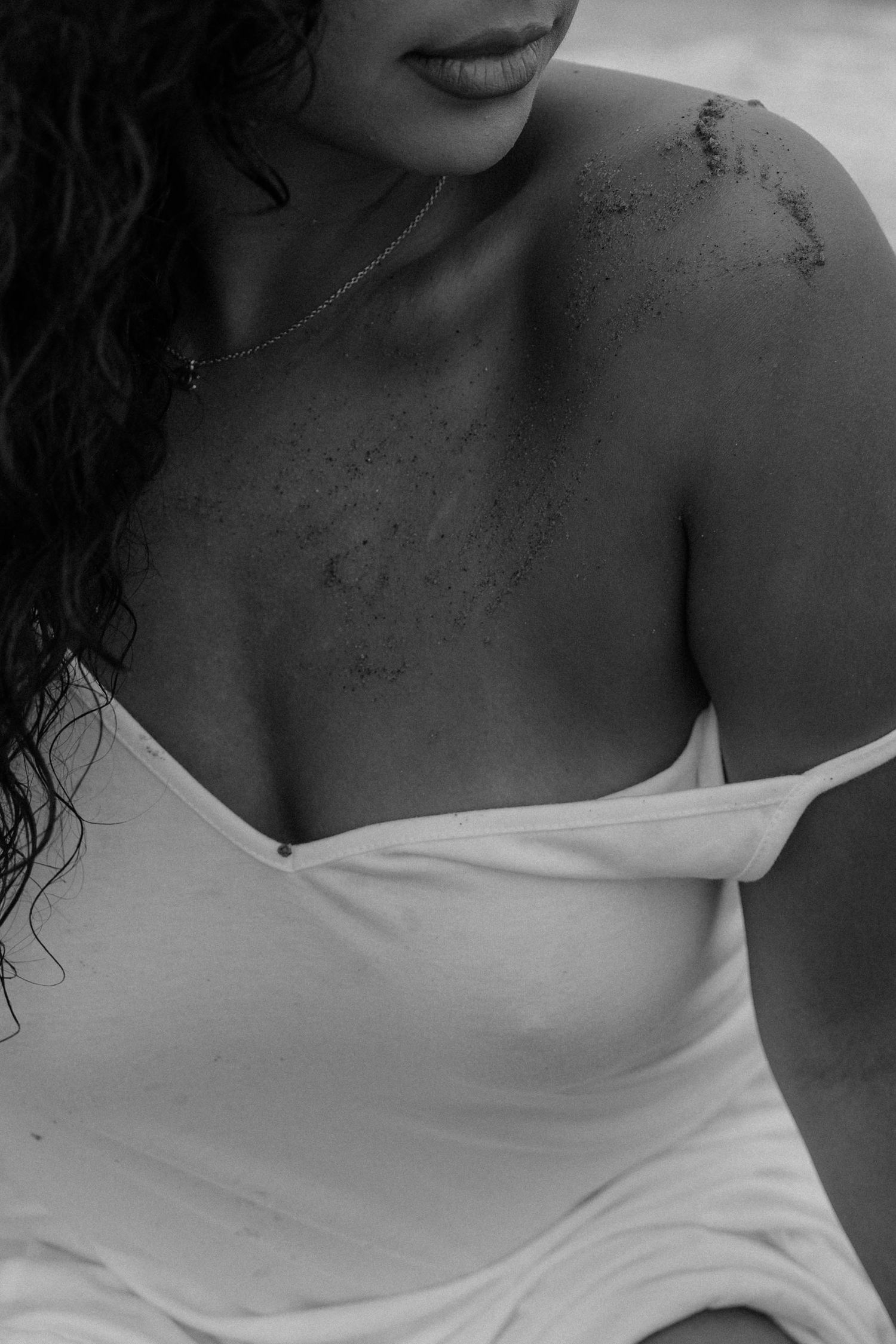 Black and white photo of a woman in a white off-shoulder dress with sand on her shoulder at the beach.