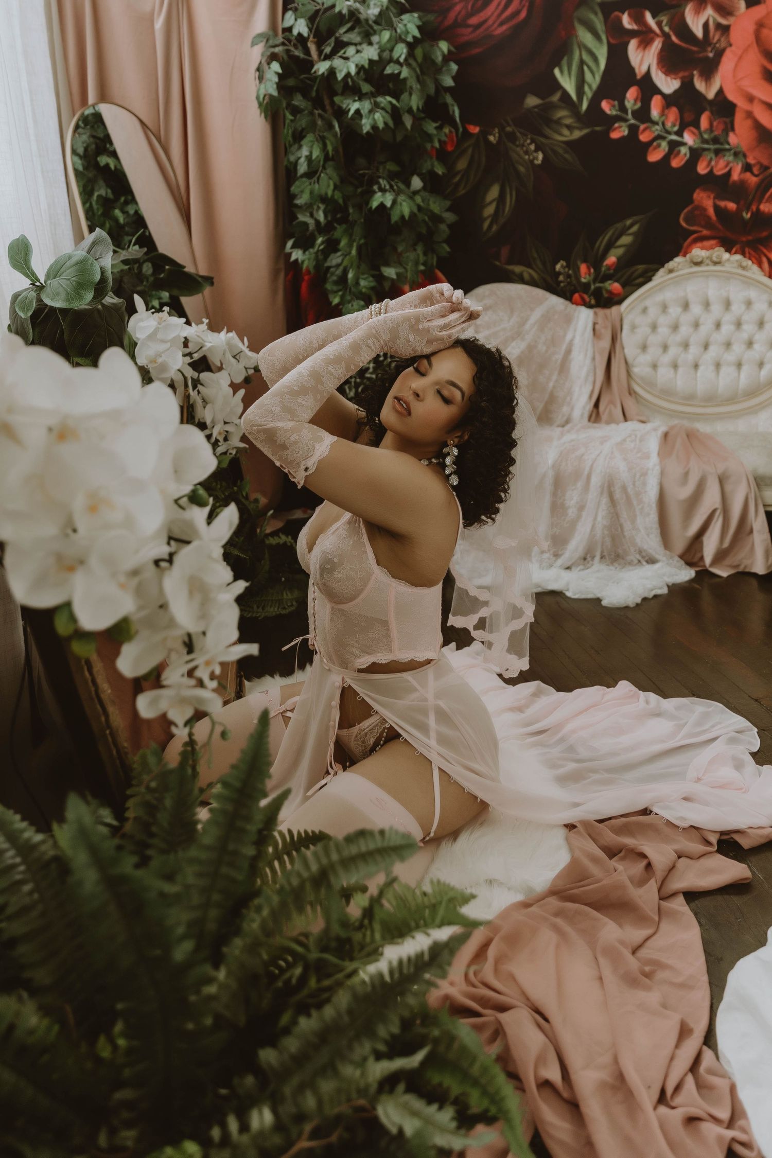 Woman in white lace lingerie poses gracefully on floor surrounded by lush greenery and white orchids in elegant floral room.