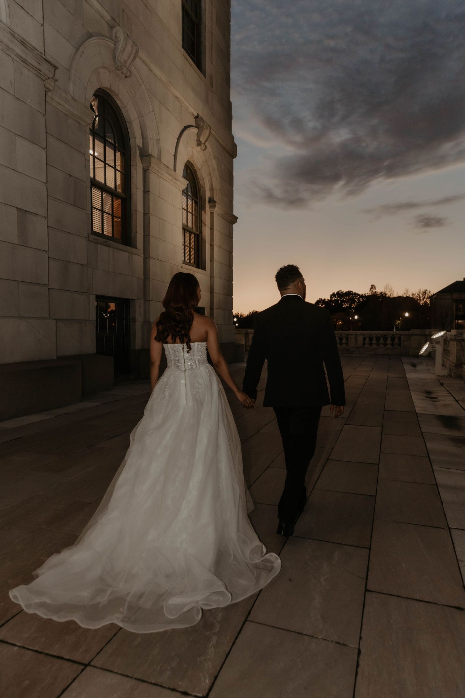 Bride in a white tulle gown and groom in a dark suit hold hands walking along a stone terrace at dusk past a historic building.