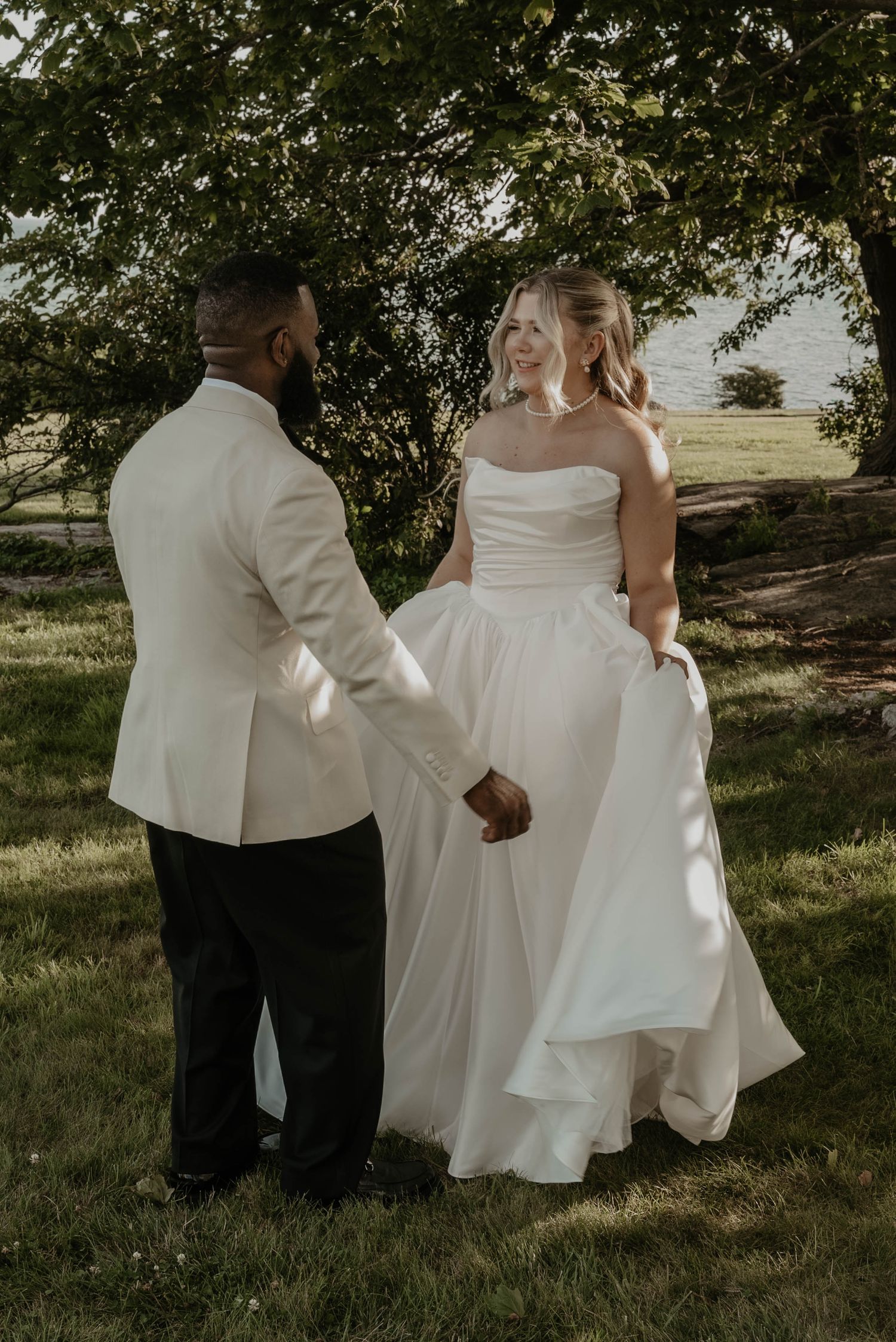 Bride in a strapless white satin gown holds hands with a groom in a cream jacket while smiling at each other under trees by the water.