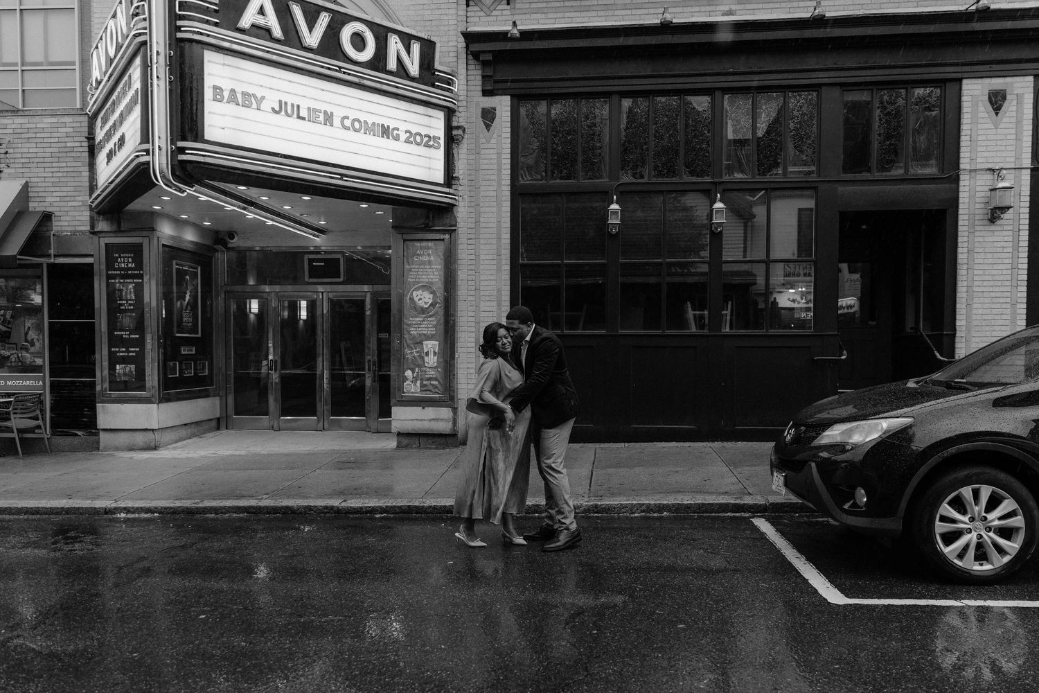 Couple embraces outside Avon Theatre with Baby Julien Coming 2022 marquee in black and white photo.