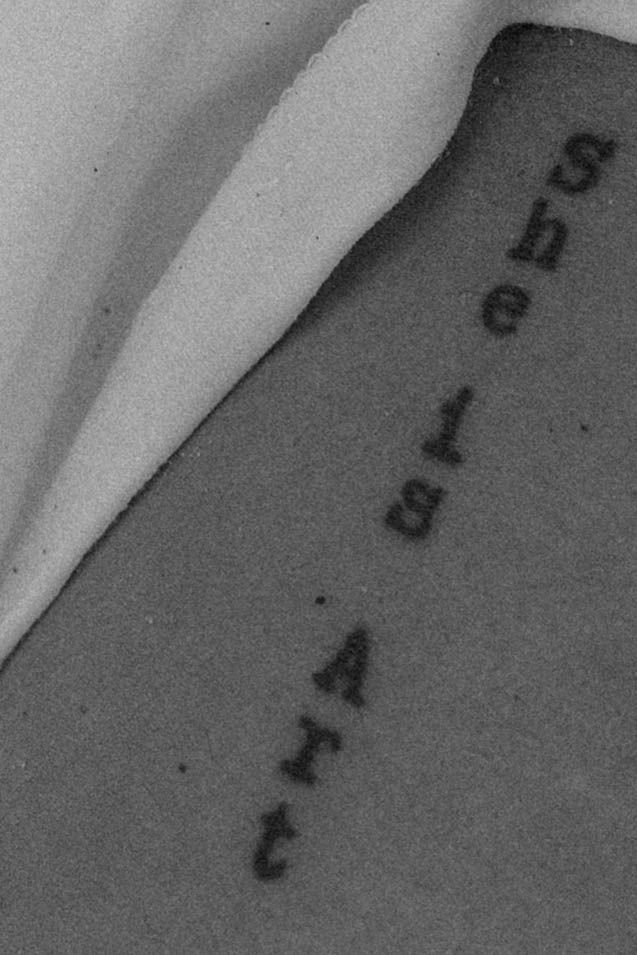 Black and white photo of a tattoo reading 'She is Art' written vertically on skin.