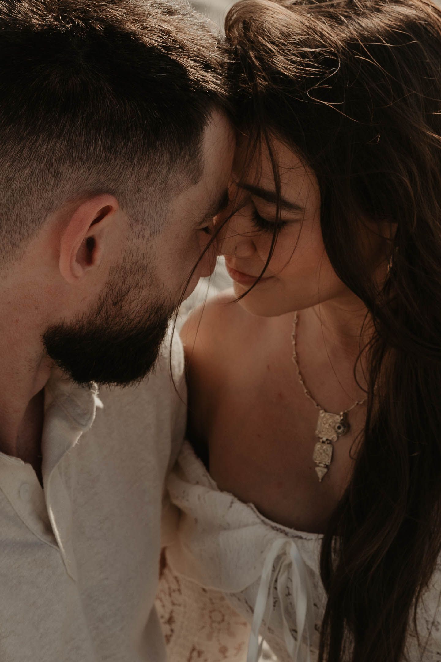 Romantic couple sharing an intimate moment, foreheads touching, woman wearing a delicate necklace in soft warm light.