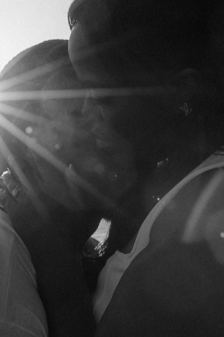 Black and white photo of couple sharing romantic kiss with sun flare glowing between them outdoors.