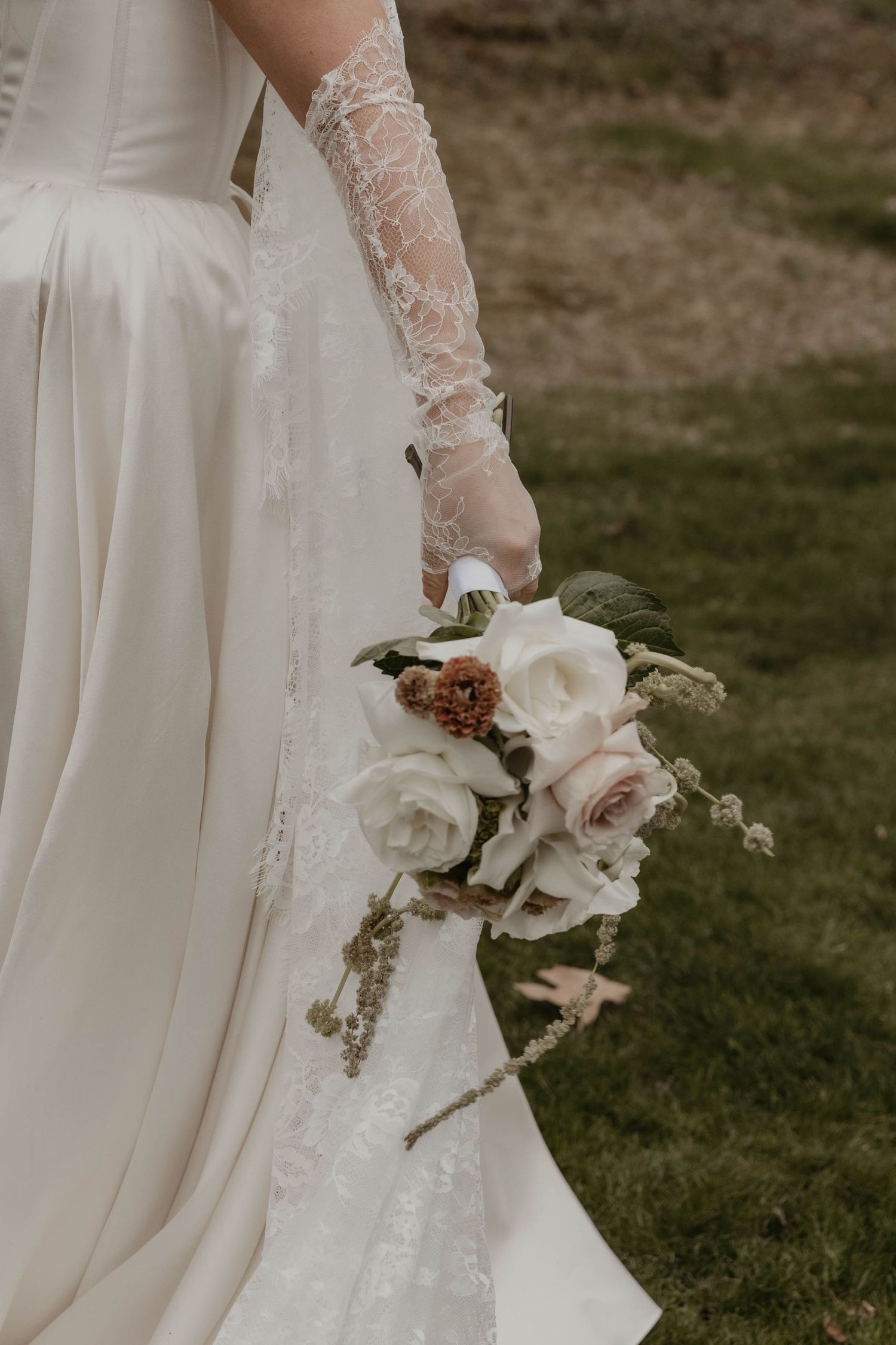 Bride in white satin gown and lace gloves holding elegant bouquet of white and blush roses outdoors.