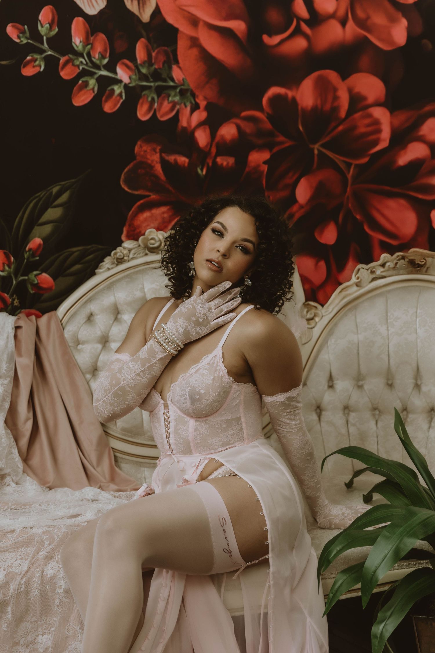 Woman in white lace lingerie and gloves poses elegantly on a vintage sofa before a bold floral backdrop.