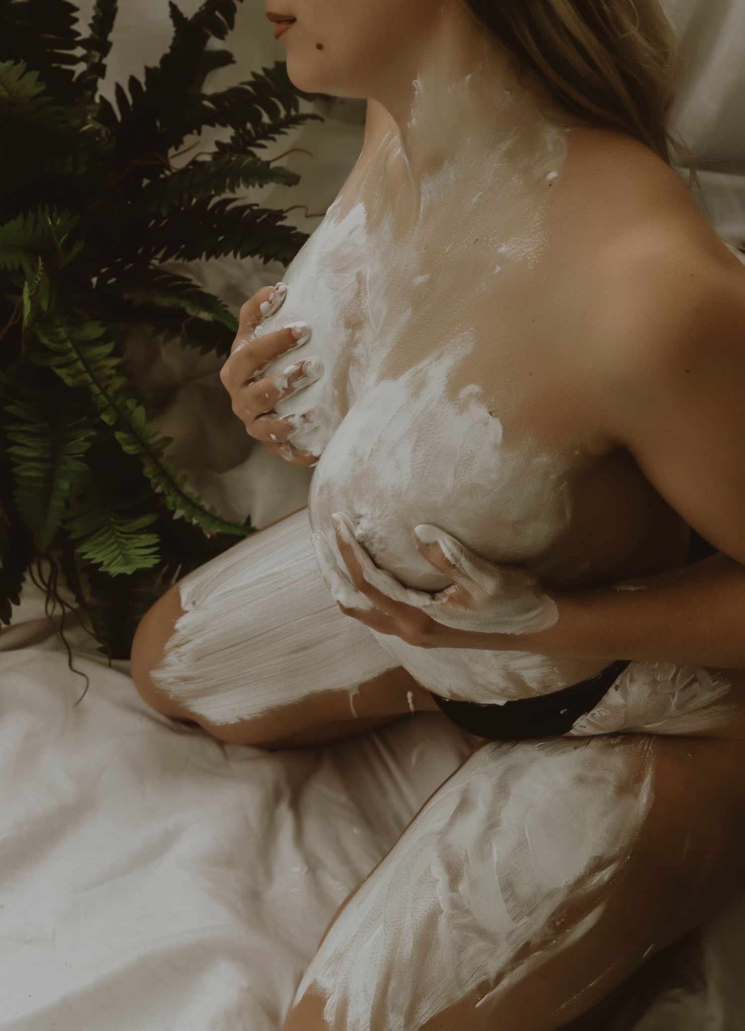 Woman kneeling on white bedding with white cream or lotion smoothed over her body, beside a green fern in soft natural light.