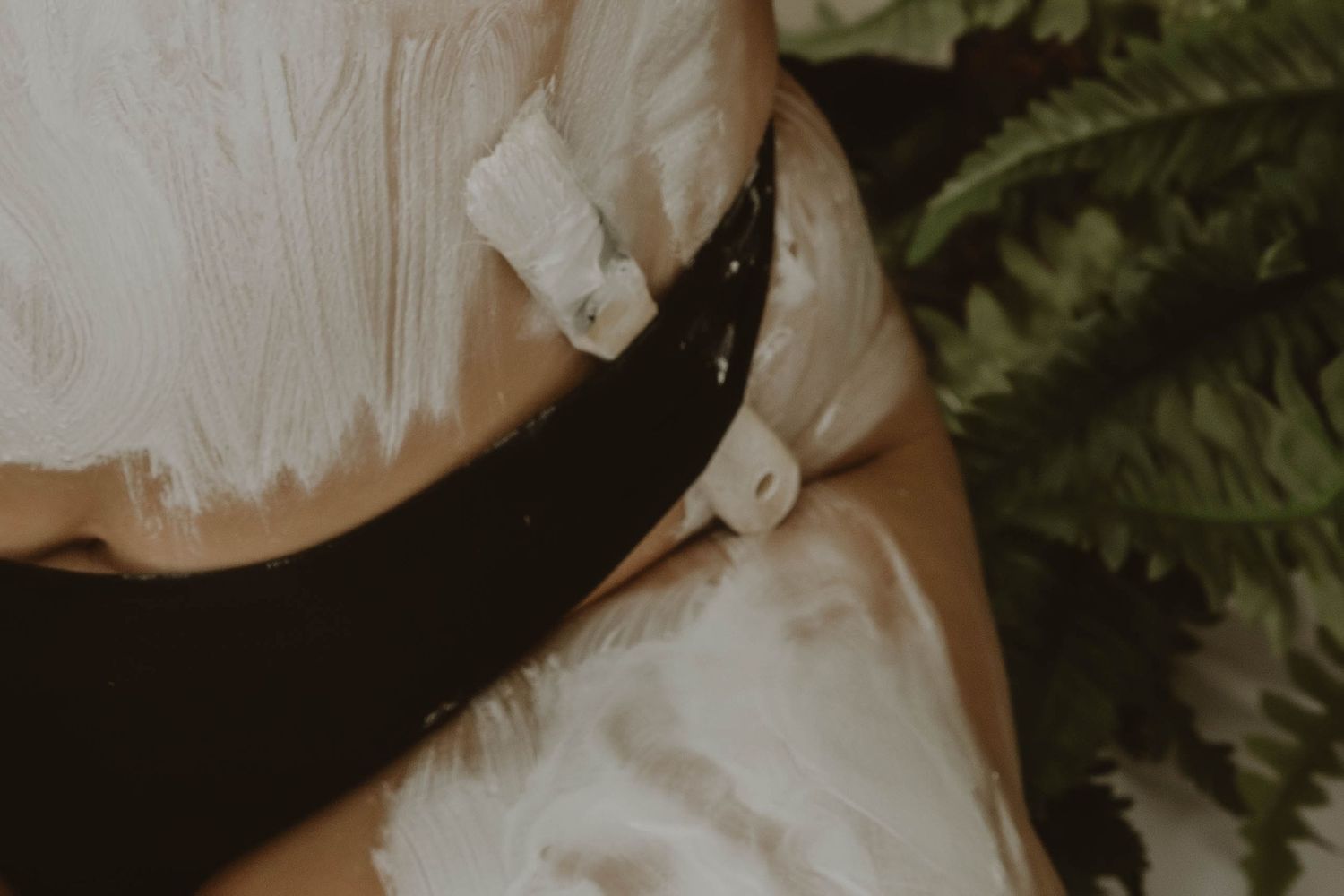 Close-up of a person's torso covered in thick white cream or foam, with a black waistband tied at the sides beside green fern leaves.