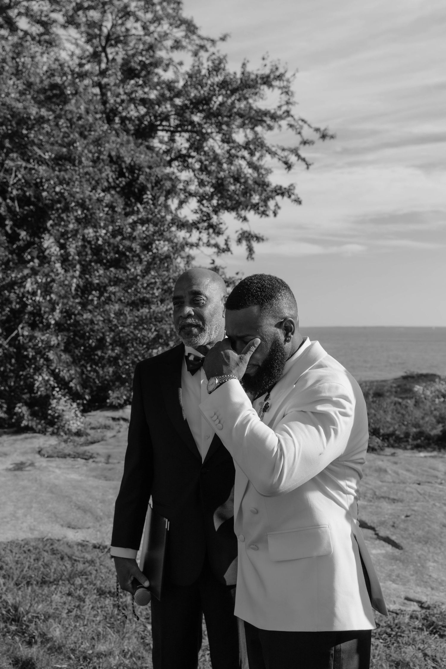 Black and white photo of two men in formal suits sharing an emotional moment outdoors near the ocean.