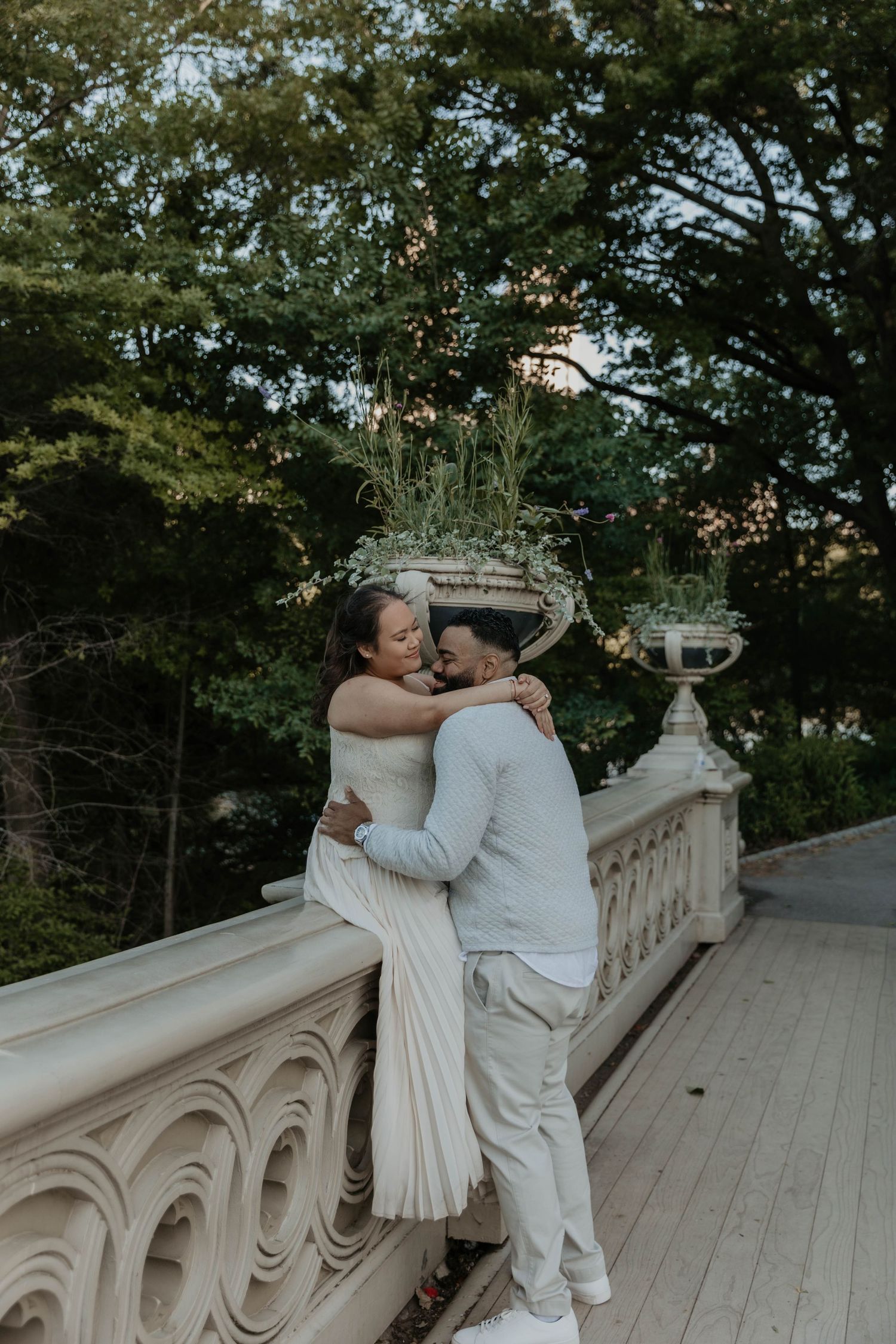 Couple embracing on ornate stone balustrade in Central Park, surrounded by lush greenery, dressed in elegant white attire.