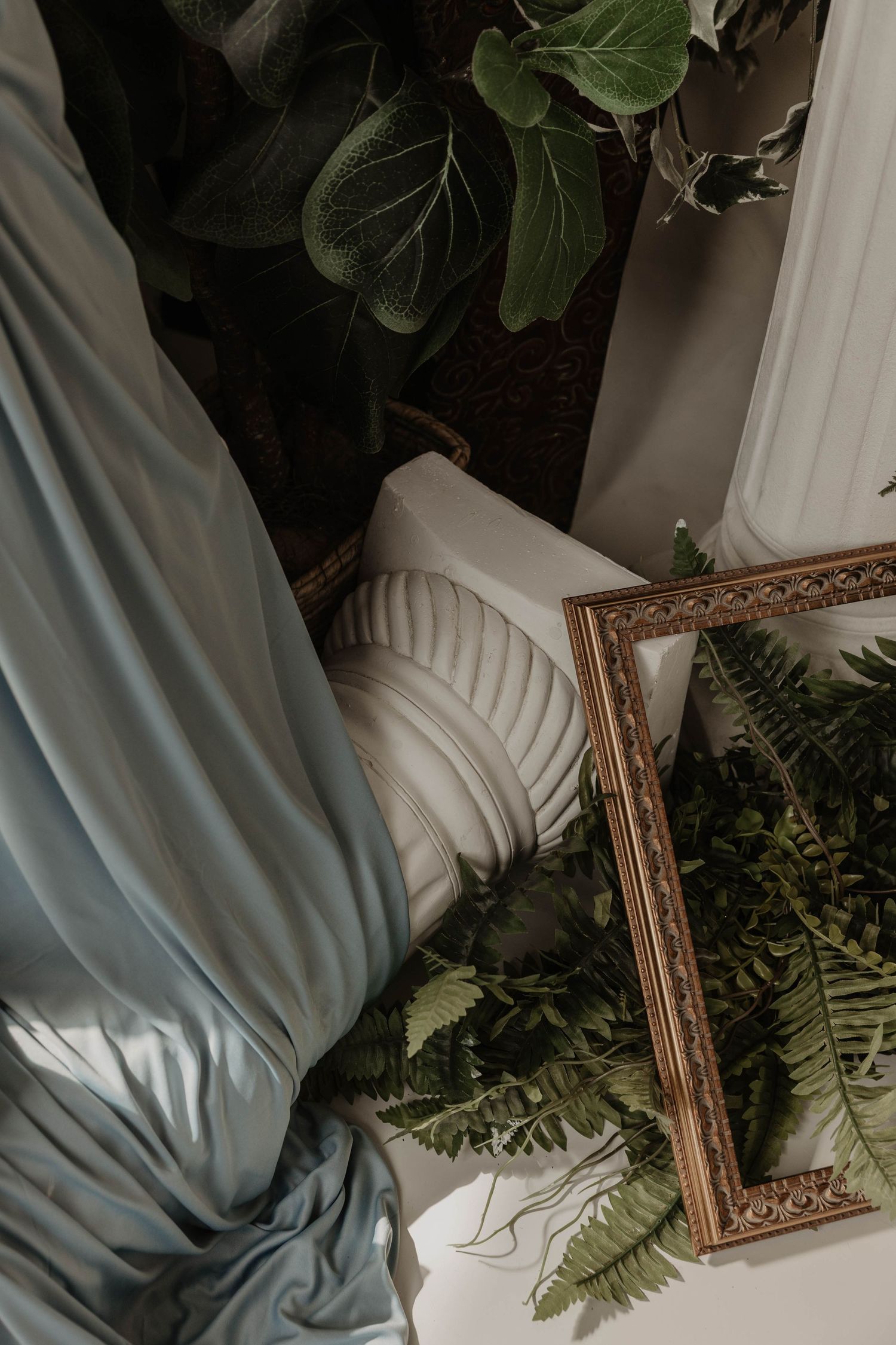 Styled vignette with cream tufted chair, gold ornate frame, ferns, and blue draped fabric near white column.