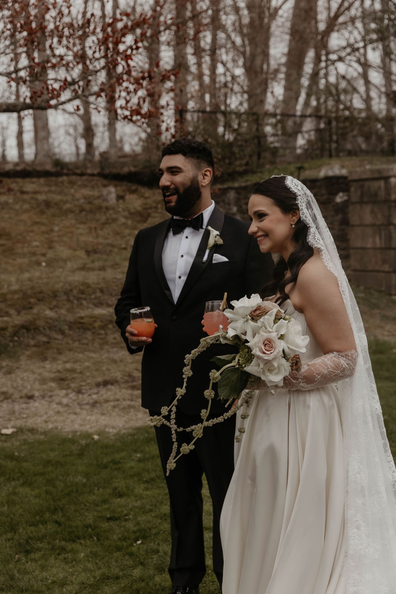 A smiling bride in a white gown with veil holds a bouquet beside a tuxedo-clad groom holding drinks outdoors.
