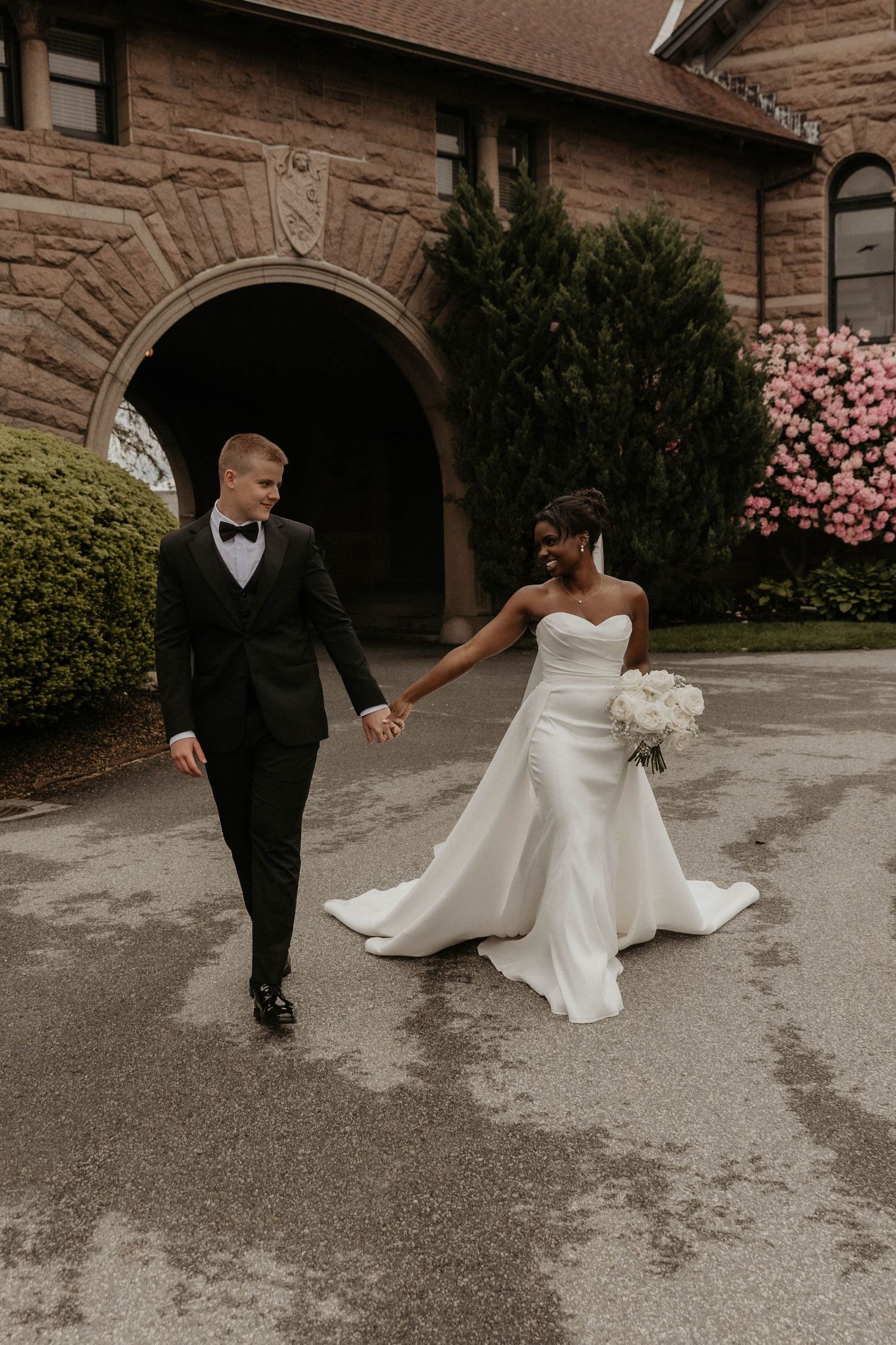 Bride and groom holding hands walking outside a stone church, bride in strapless white gown holding bouquet.