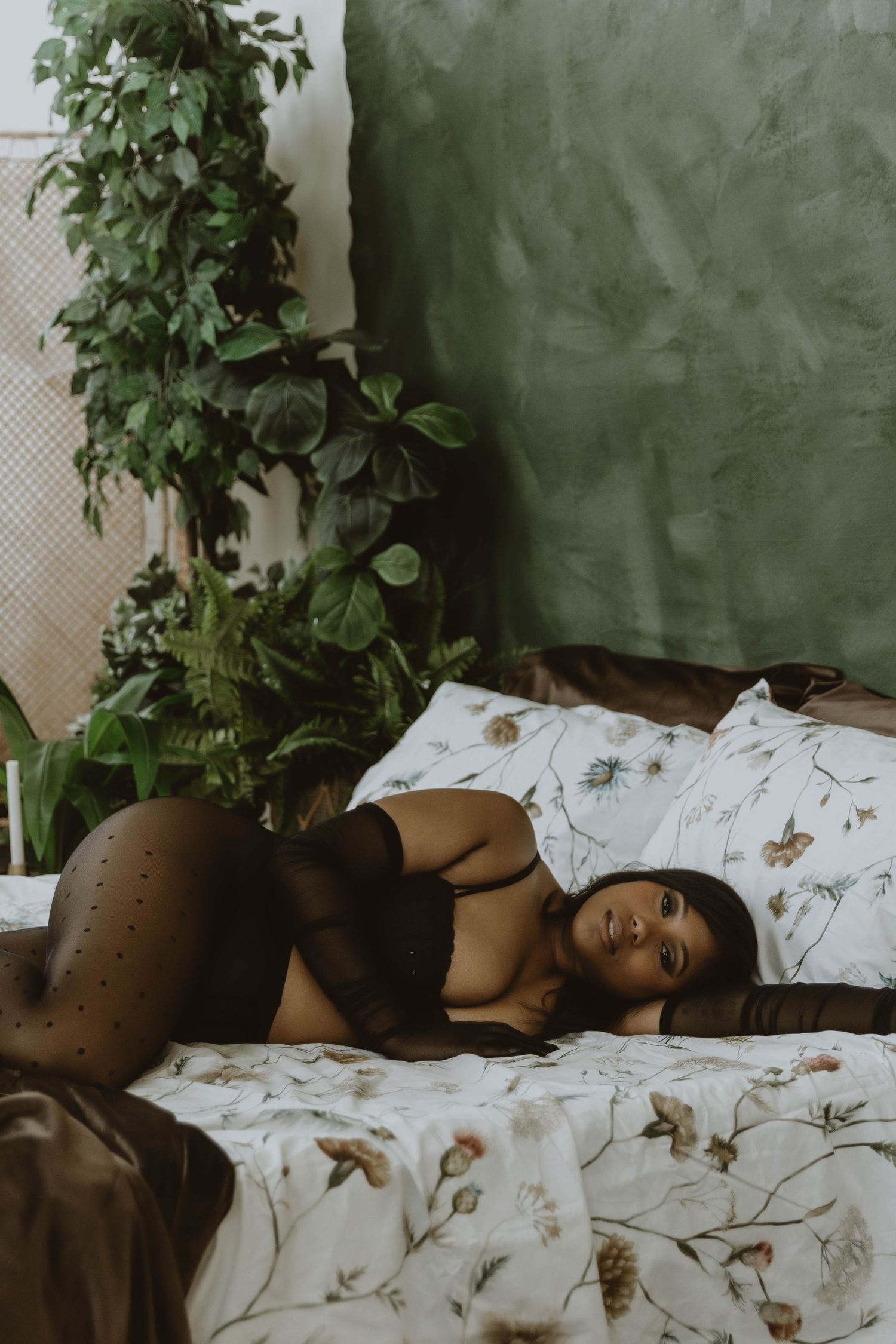 Woman in black lingerie and tights posing on a floral bed surrounded by lush green plants.
