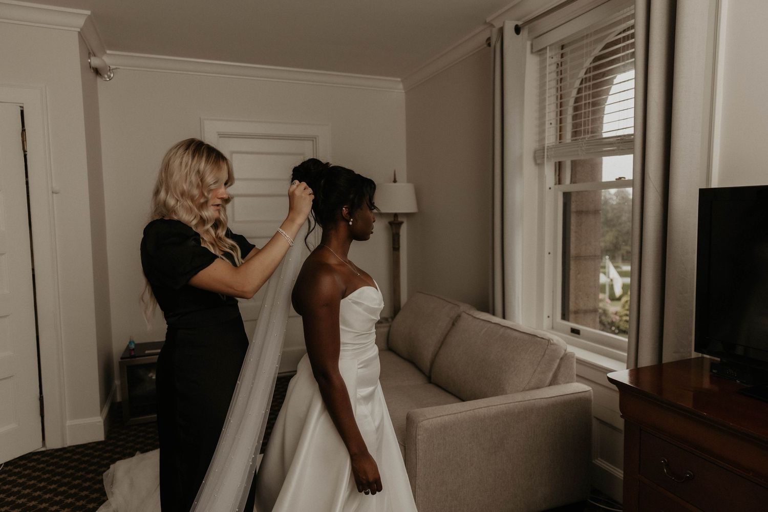 A woman in a black dress adjusts the veil of a bride in a white gown as they prepare for the wedding in a bright room.