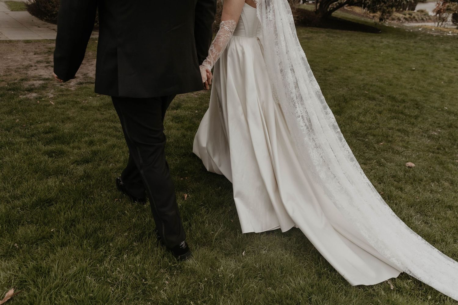 Bride and groom walking hand in hand on green grass, showcasing elegant white wedding gown with long train.