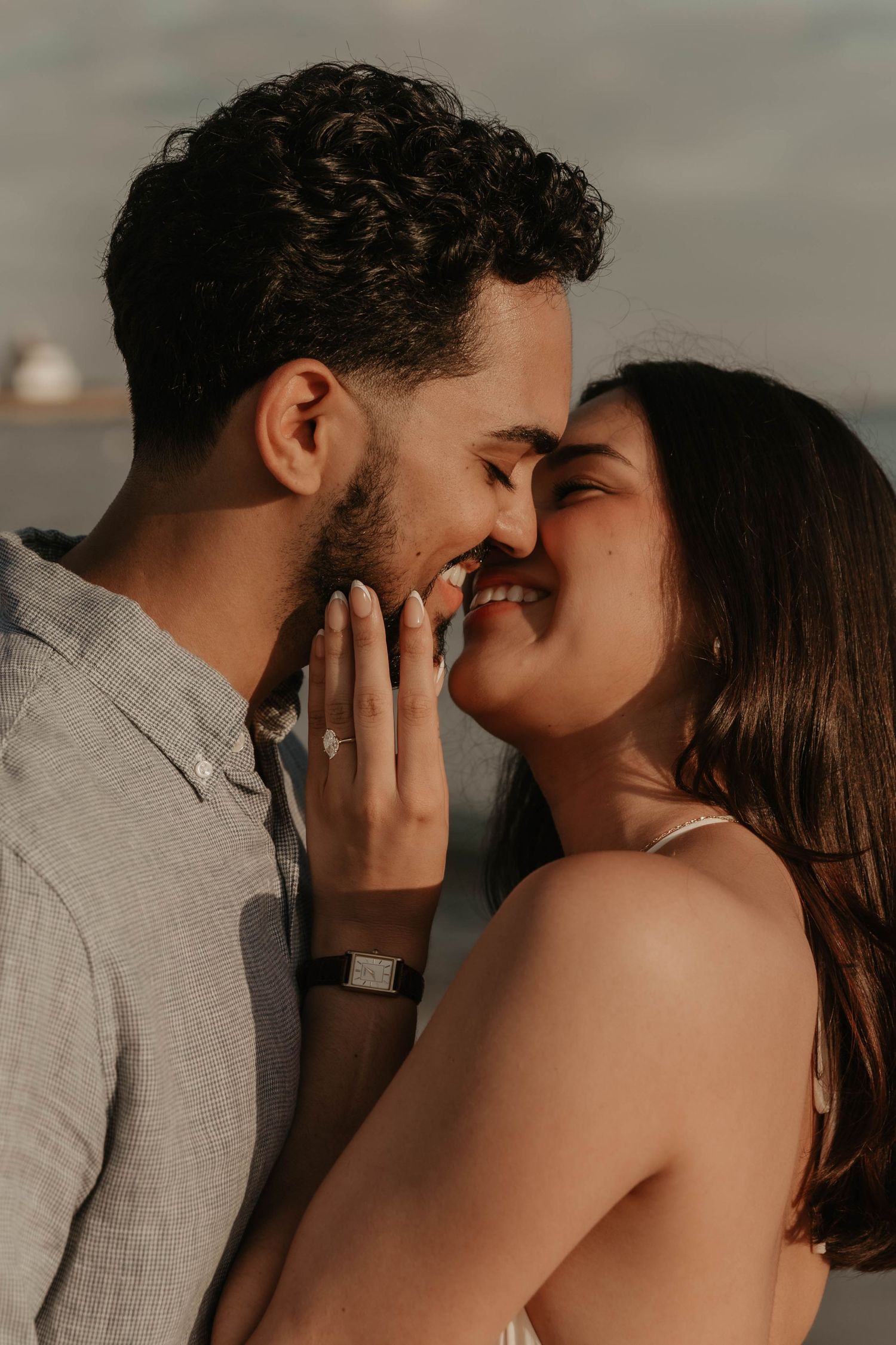 Romantic couple sharing an intimate moment, smiling nose-to-nose outdoors, woman displaying engagement ring.
