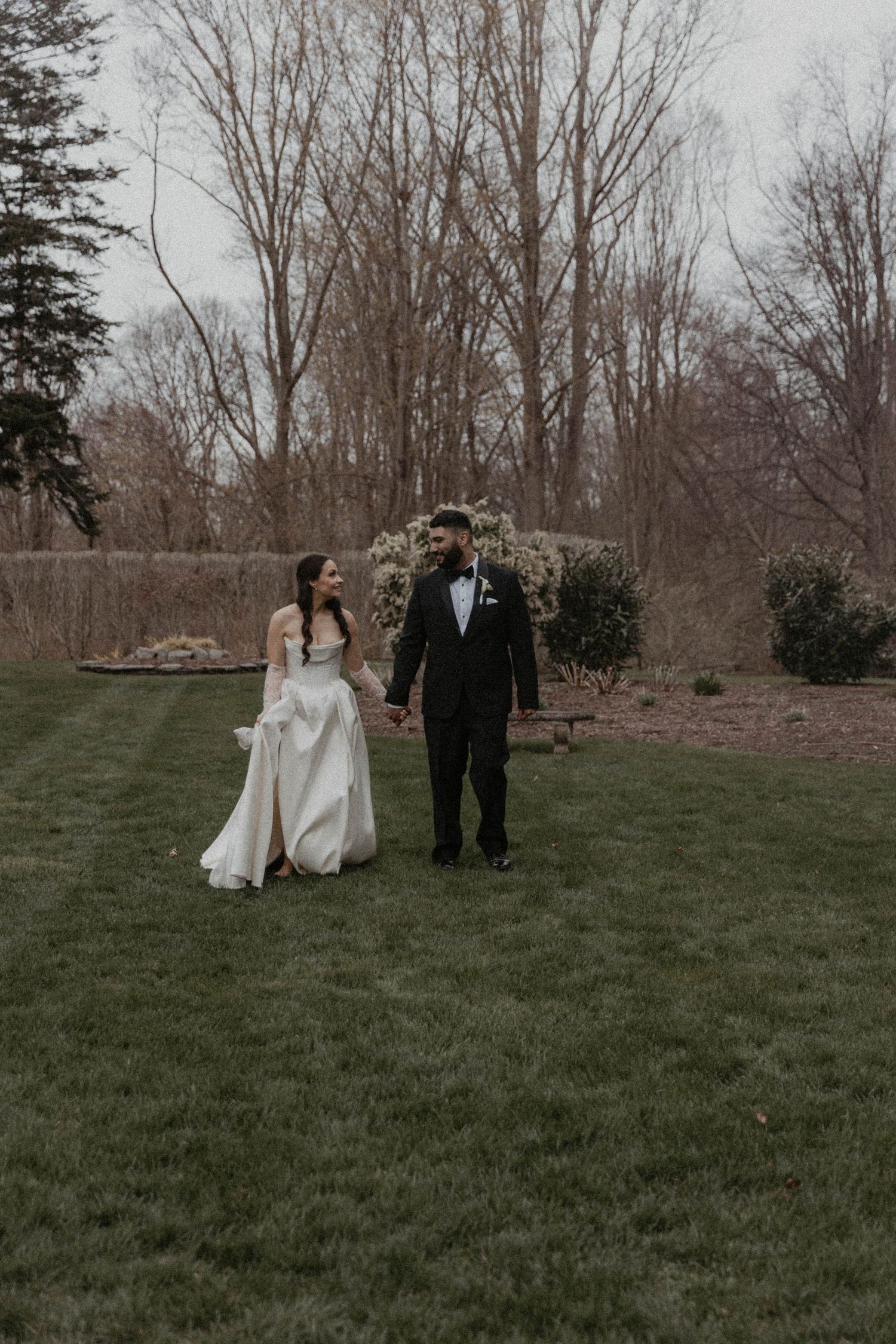5mm film photographer near Bride and groom walk hand-in-hand on lush green lawn surrounded by bare trees during outdoor wedding celebration.