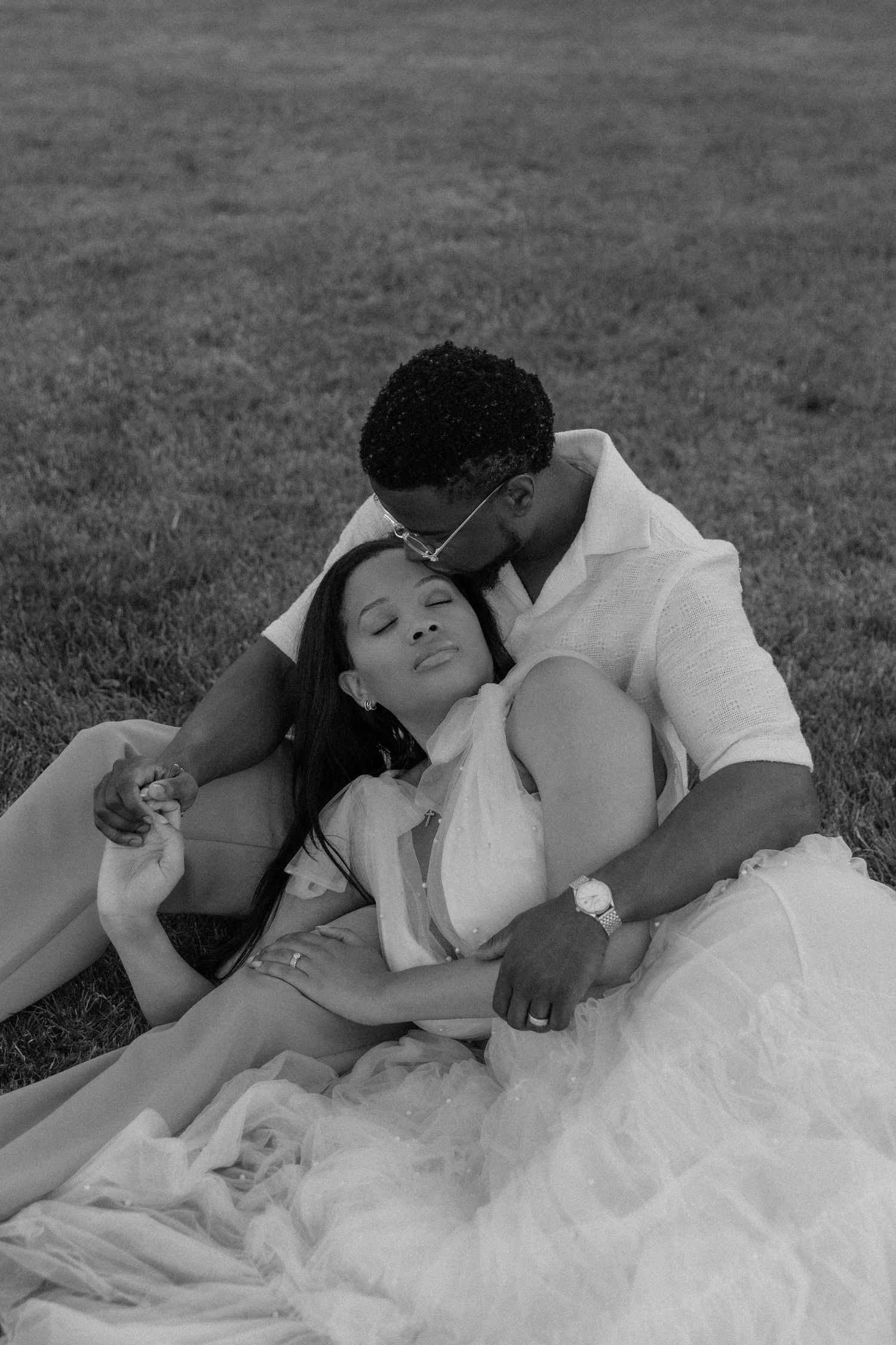 Black and white photo of a couple sitting on grass, man kissing woman on cheek during romantic outdoor session.