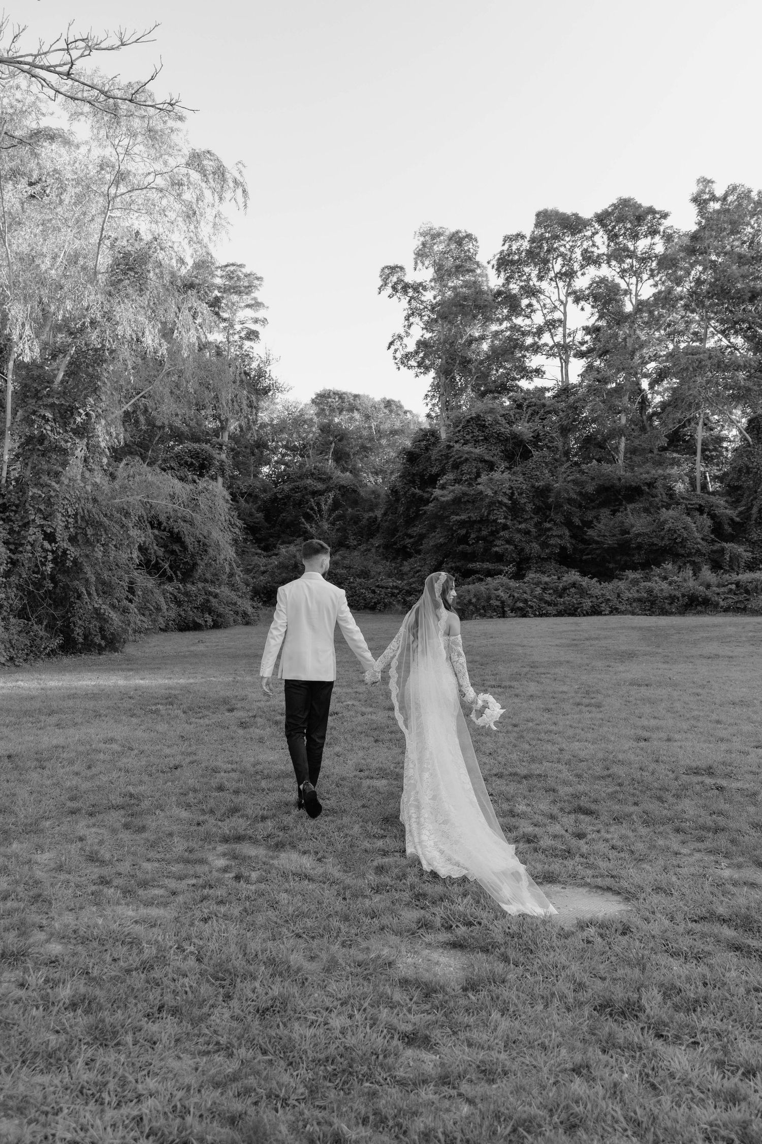 Black and white photo of a bride and groom holding hands and walking across a grassy lawn toward trees, her long veil trailing behind.