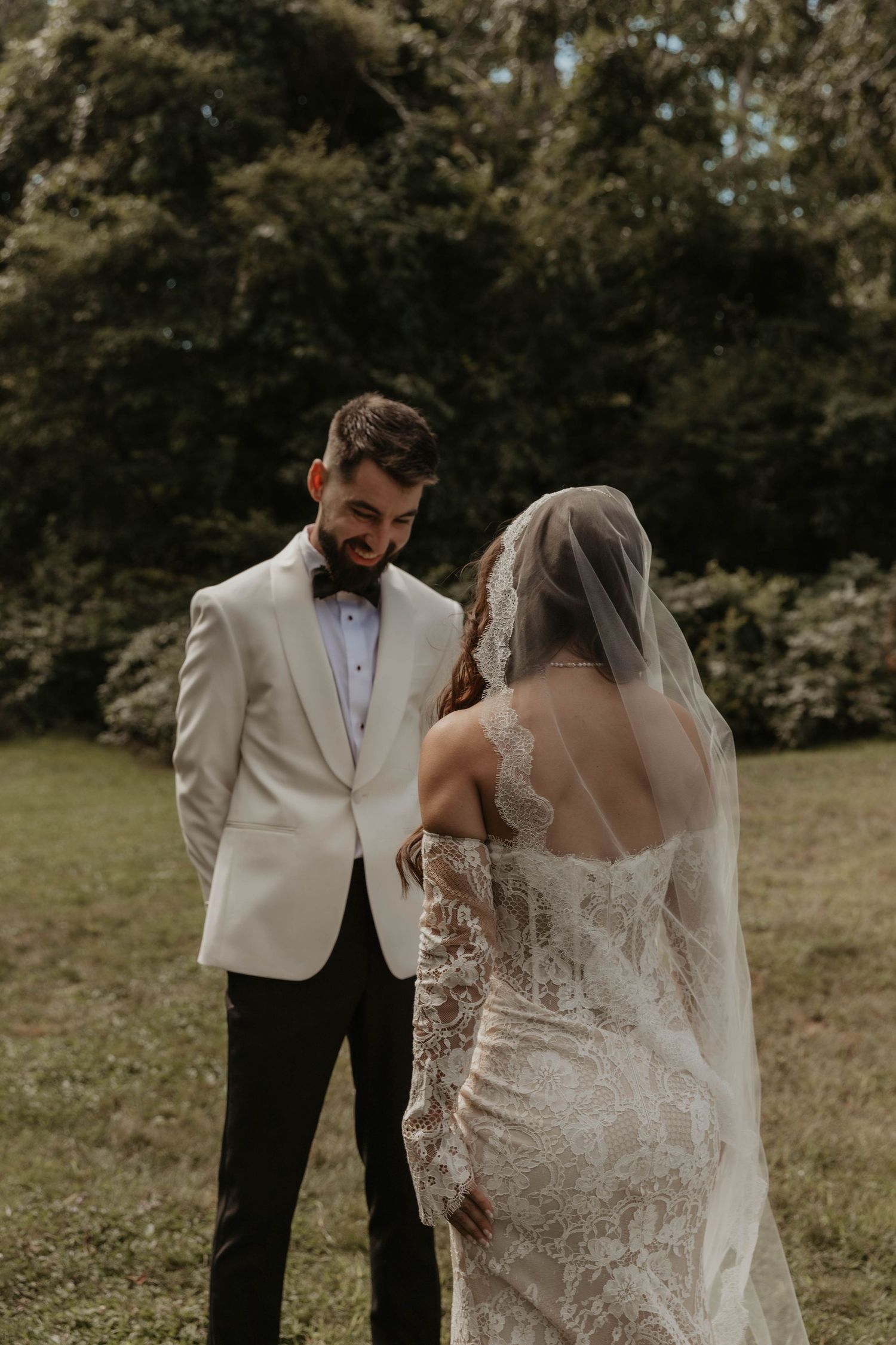 Smiling groom in a white tuxedo jacket and bow tie looks at his bride in a lace gown and veil during a outdoor first look.