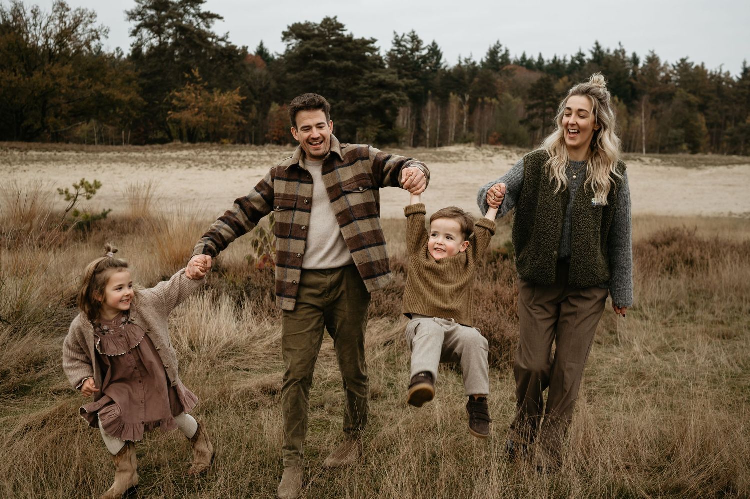 Een spontaan gezin speelt in het hoge gras tijdens een herfstfotoshoot in een rustieke buitenomgeving.