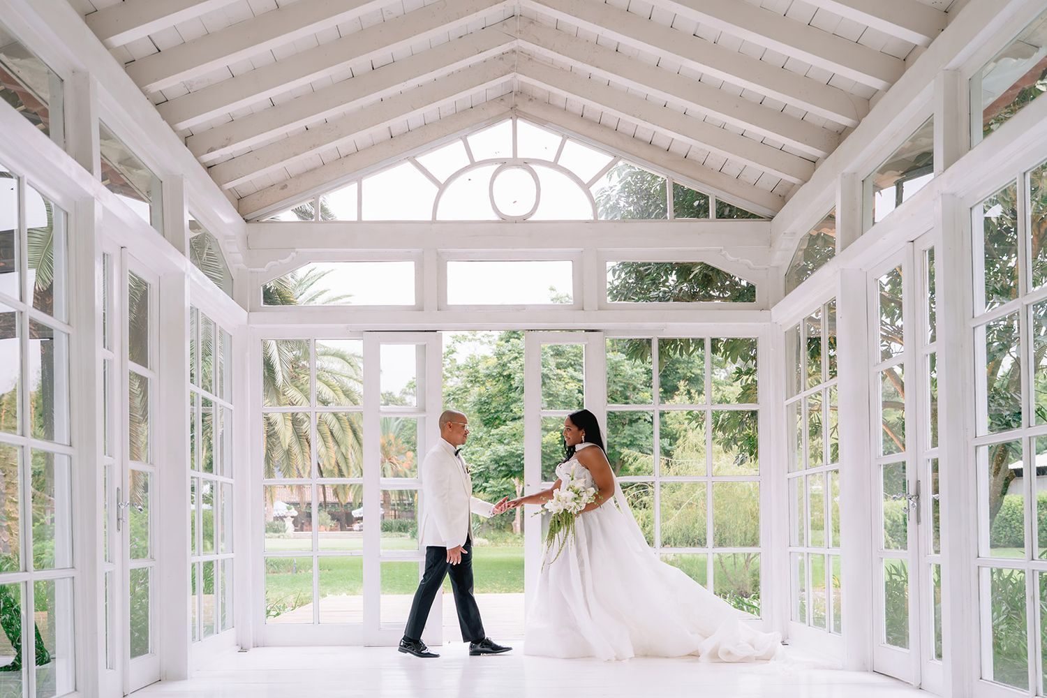 Bride and groom hold hands in a stunning white glass conservatory with garden views on their wedding day.